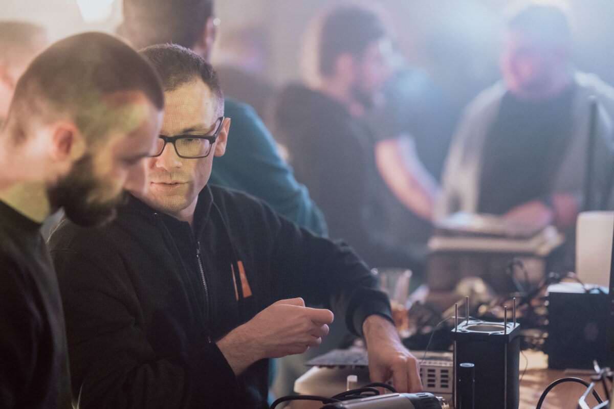 Two men are having a discussion at a table set with appliances and electronic equipment. The man on the right, wearing glasses, points to something on the table. The foggy background reveals more people and activity in the dimly lit room, capturing the essence of the event photo essay.  