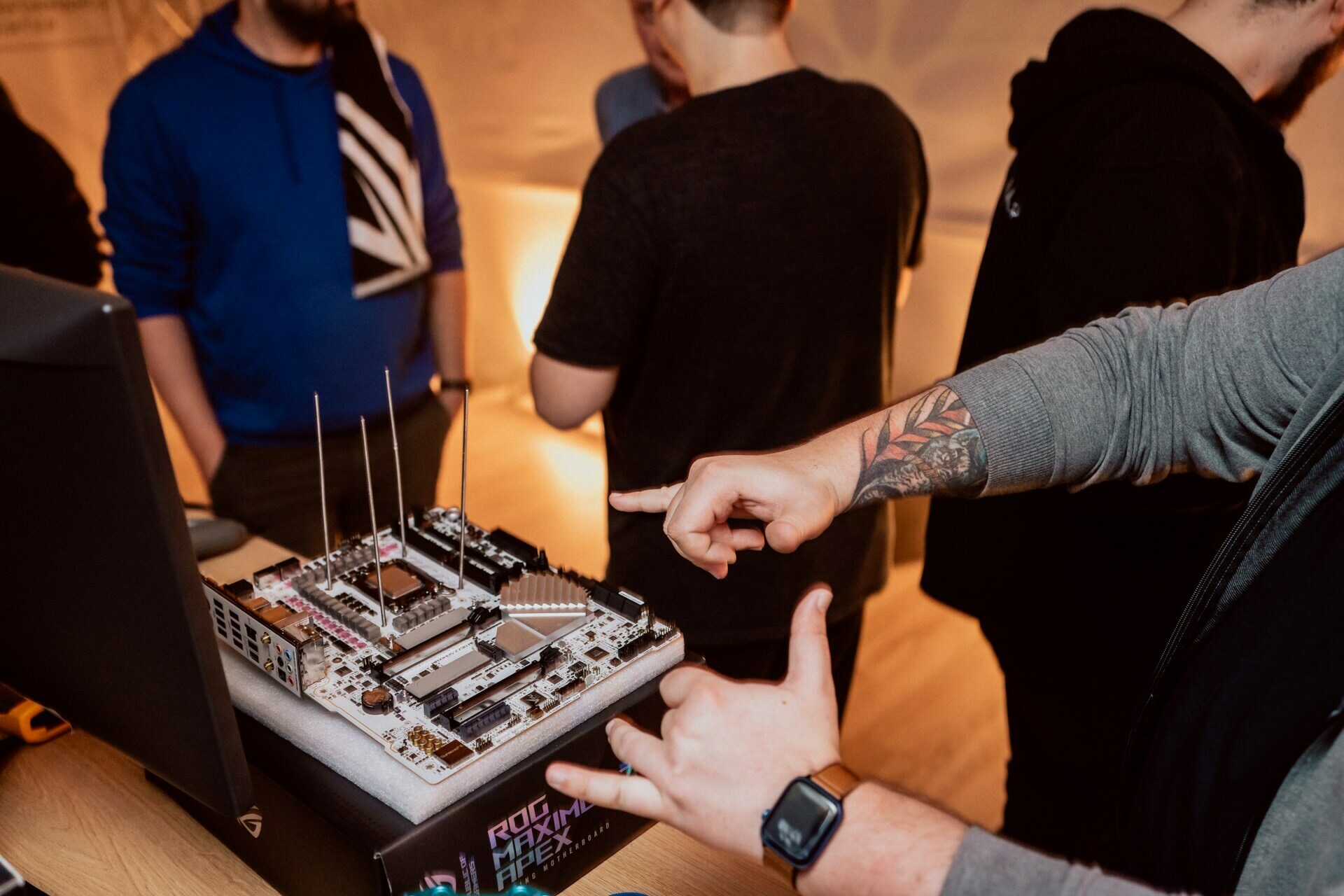 A person with a tattooed arm points to a circuit board on a table, surrounded by computer equipment. Three other people stand in the background, talking. The scene appears to be a simple technical workshop or meeting, perfect for an event photo shoot or anyone looking for an event photographer warsaw.  