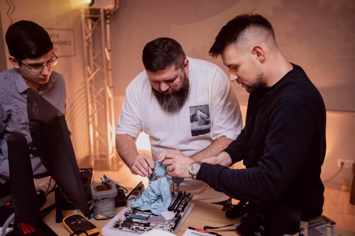 Three people gather around a table and work on assembling or repairing a computer. One person with a beard holds a rag, another helps. The third person watches closely. The table is swamped with various components and computer tools that capture the essence of event photography warsaw.   