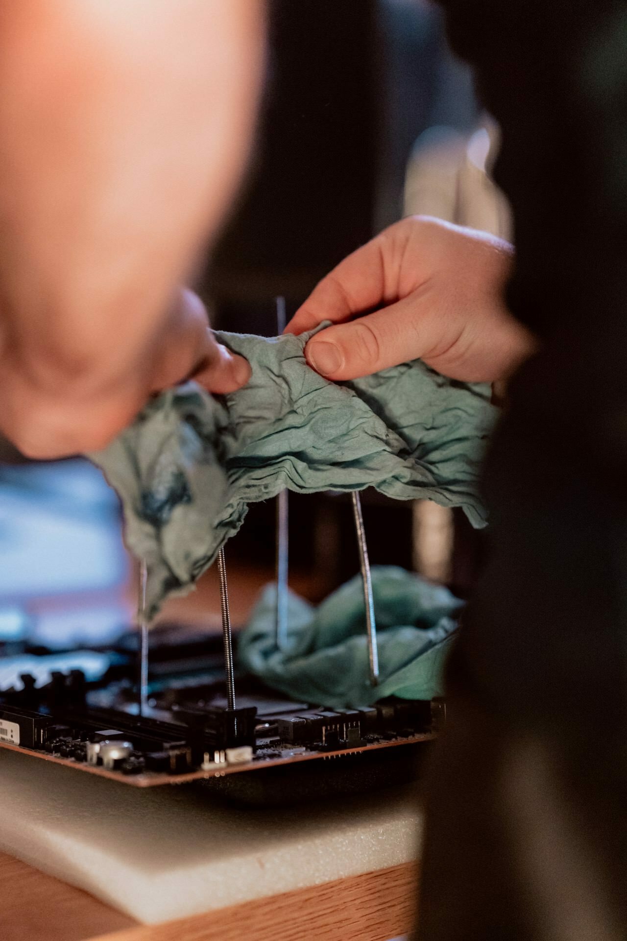 A person uses a soft cloth to wipe or clean a computer motherboard laid out on a table. The figure can be seen from the shoulders down, focusing on the hands holding the cloth and the electronic components on the board, creating a detailed photo report of the event. 