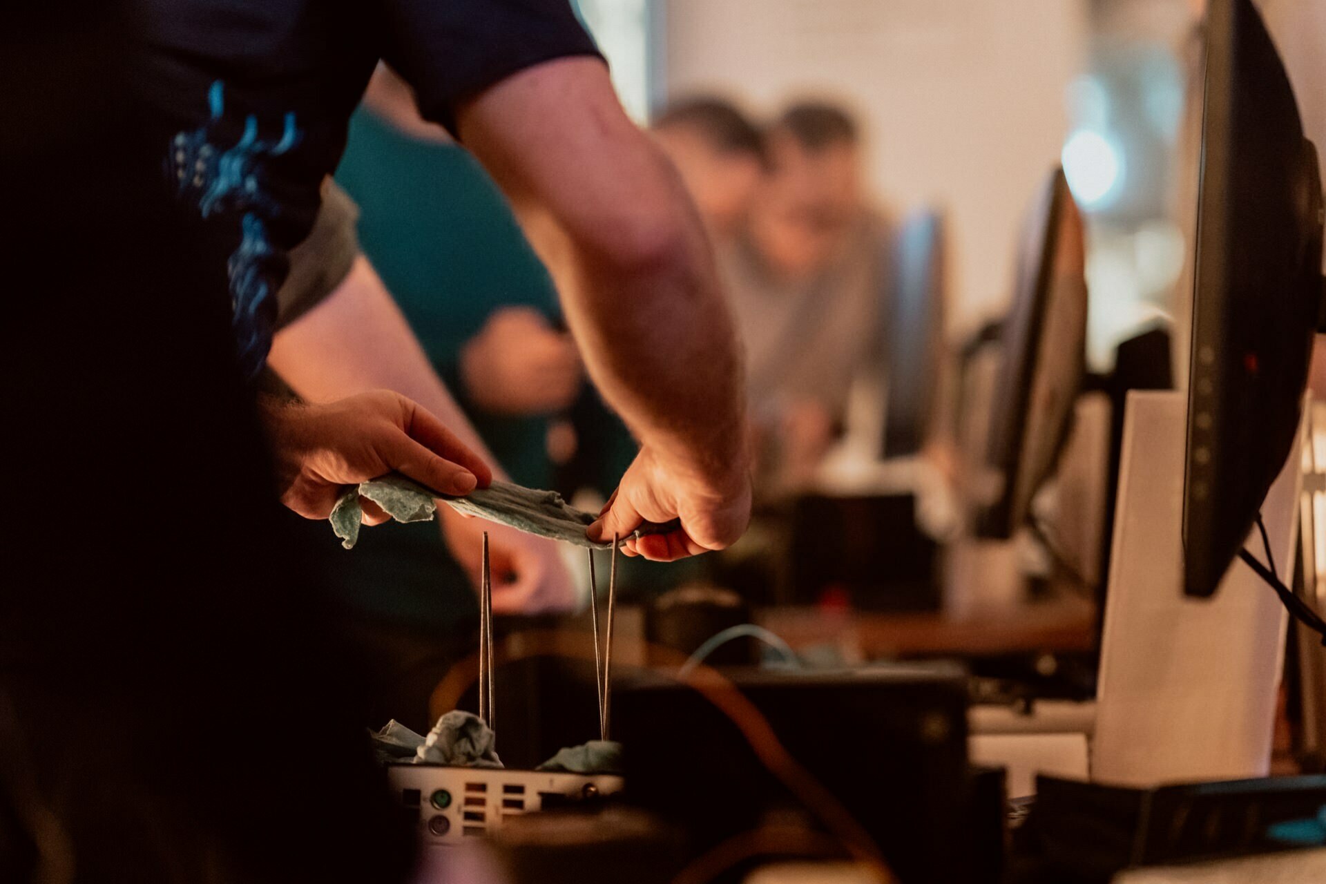 People work with various electronic devices and monitors, performing tasks with their hands visible. The focus is on the hands of people holding and stretching material, such as string or wire, in busy indoor environments. This scene could be part of an event photography showing intricate details.  