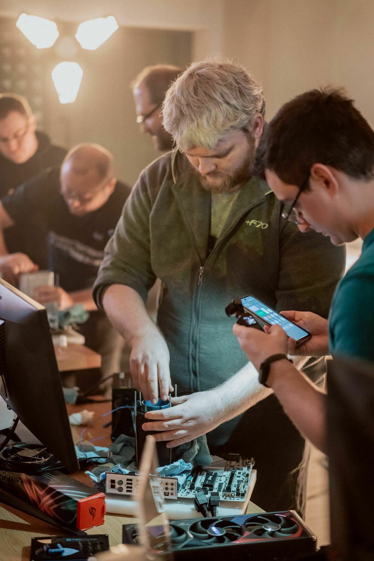 A group of people are working together to assemble or repair computer components at a table. One person in the foreground is concentrating on putting the parts together, while another is holding a smartphone nearby and capturing a photo report of events. Various computer parts and tools are scattered on the table.  