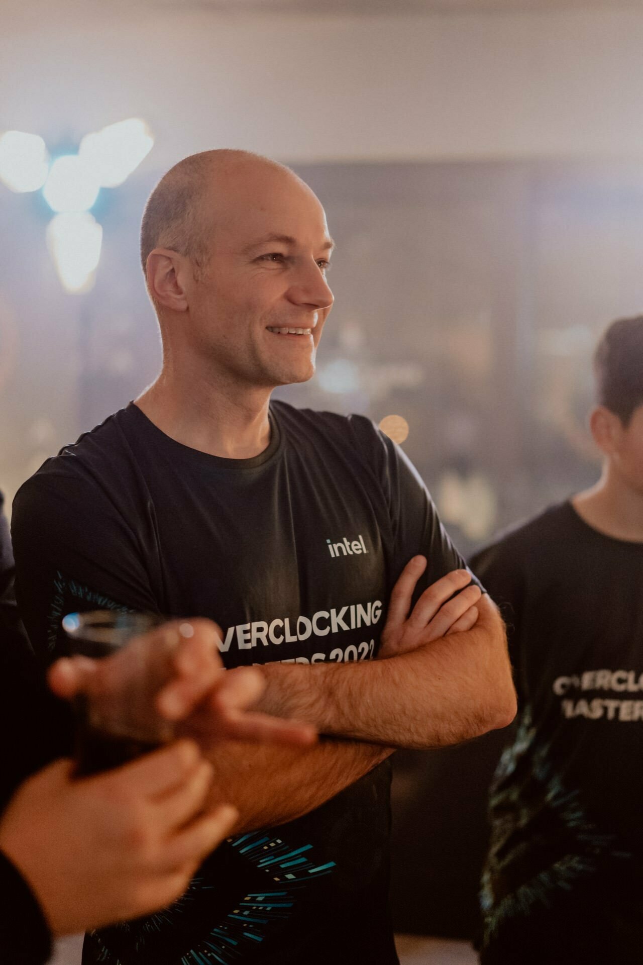 A person wearing a dark shirt with the words "Intel" and "OVERCLOCKING MASTERS 2021" is standing with his arms folded, smiling and looking to the left. They are in a room with a blurred background, and next to them is another partially visible person. This event photography captures the moment perfectly.  