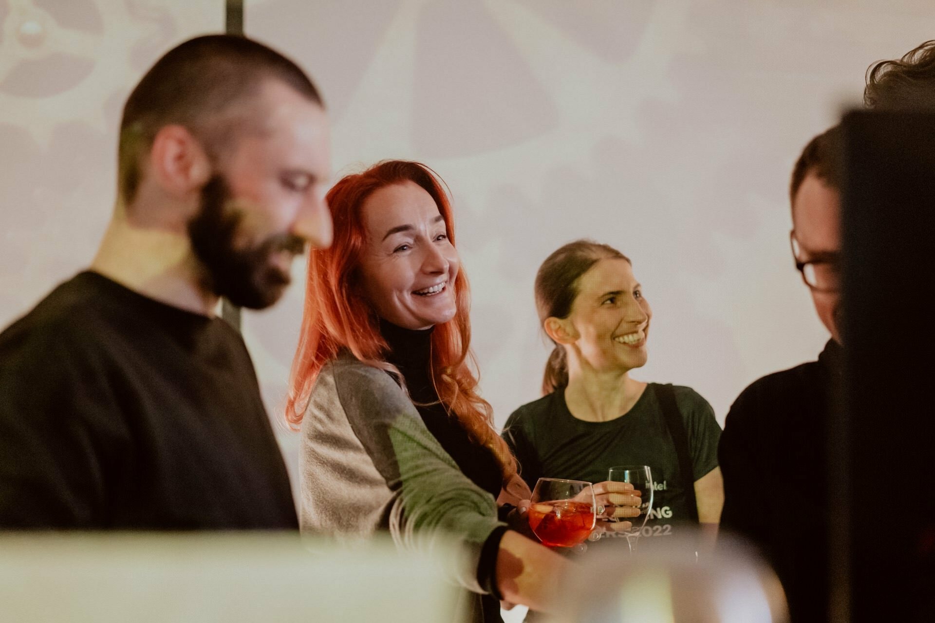 A group of four people, three women and a man, have gathered close together in a relaxed atmosphere. They smile and engage in conversation. One woman with long red hair holds a drink. The softly lit background creates an inviting atmosphere, making it ideal for event photography.   