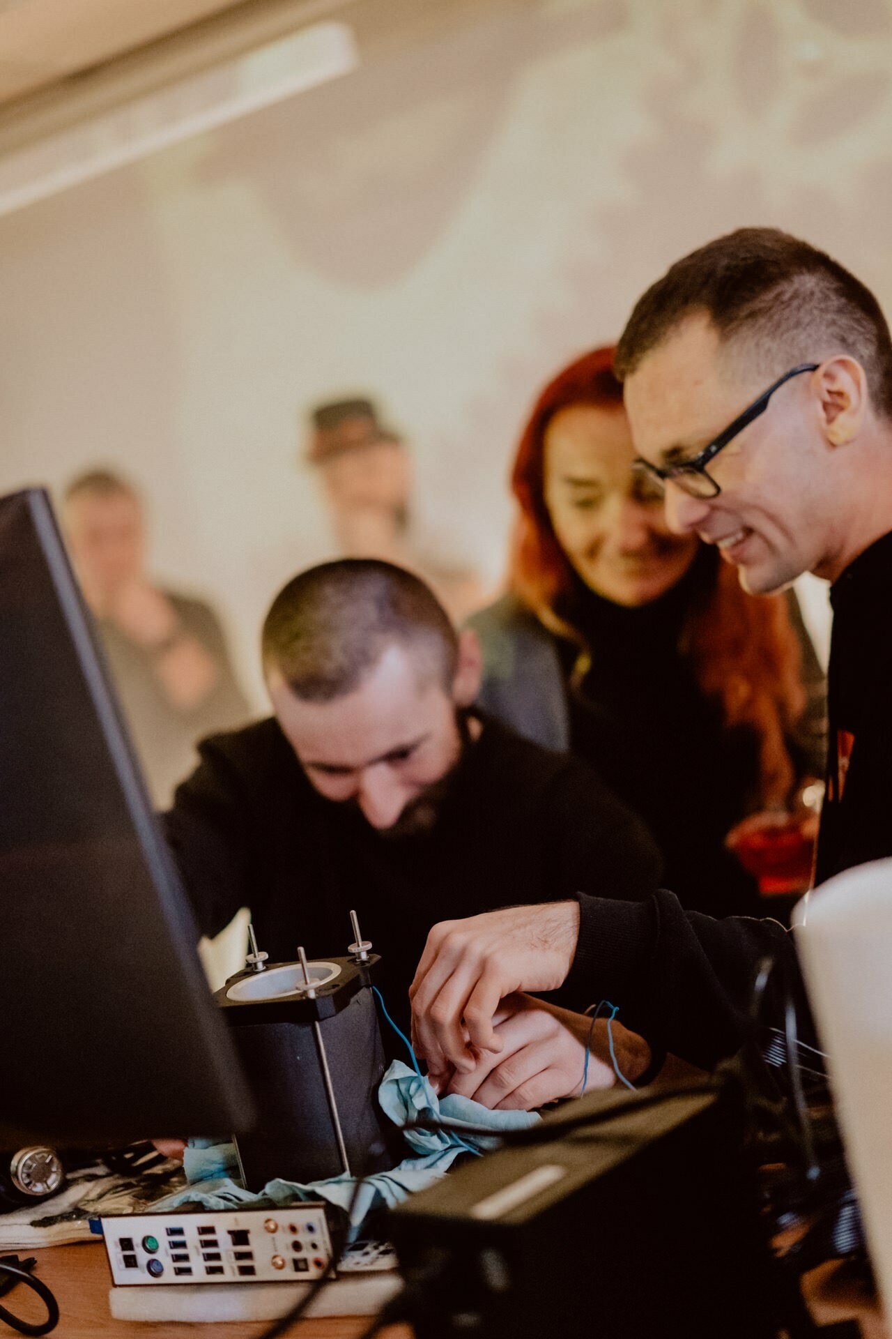 Three people gather around a table with electronic equipment and wires. One person sits and focuses on the equipment while the other two, a woman with red hair and a man with glasses, stand nearby, smiling and observing. The background is blurred, capturing the essence of event photography.  