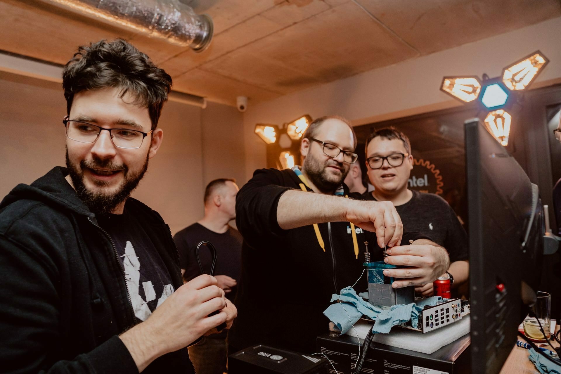 Three men gathered around a desk with computer equipment and other hardware are working together on a project. One man holds the cable while the other two focus on the equipment, and one of them adjusts the components. The environment suggests a common technological setting, ideal for a photo-op of the event.  