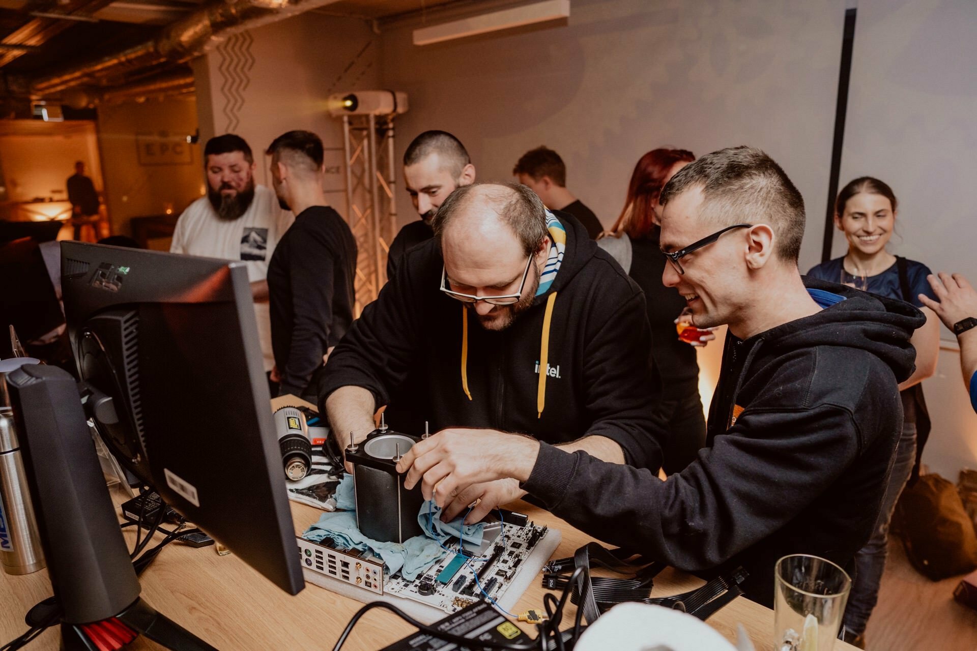 A group of people have gathered around a table with computer equipment. Two men in the foreground are assembling parts, while others are watching and talking in the background. The collaborative and focused atmosphere is beautifully captured, typical of Marcin Krokowski's event photojournalism, with tools and components scattered on the table.  