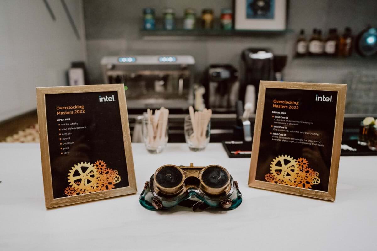 On the tabletop are two framed lists of Intel Overclocking Masters 2022 events, a set of vintage goggles, and in the background an assortment of cafe supplies, including stirrers, a coffee maker and various jars. A detailed photo recap of the events is captured in this vibrant setting. 