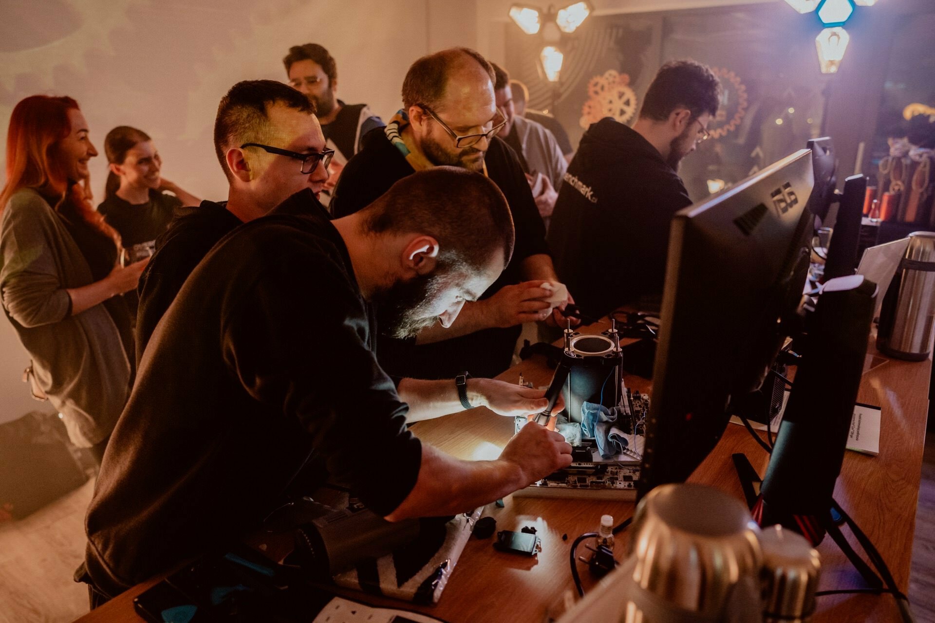 A group of people gather around a table of computers and electronic equipment. The person in the foreground is busily working on a circuit board while others watch. The room has a warm, dimly lit atmosphere with industrial decor and pinwheels on the wall, capturing the essence of event photographer warsaw.  