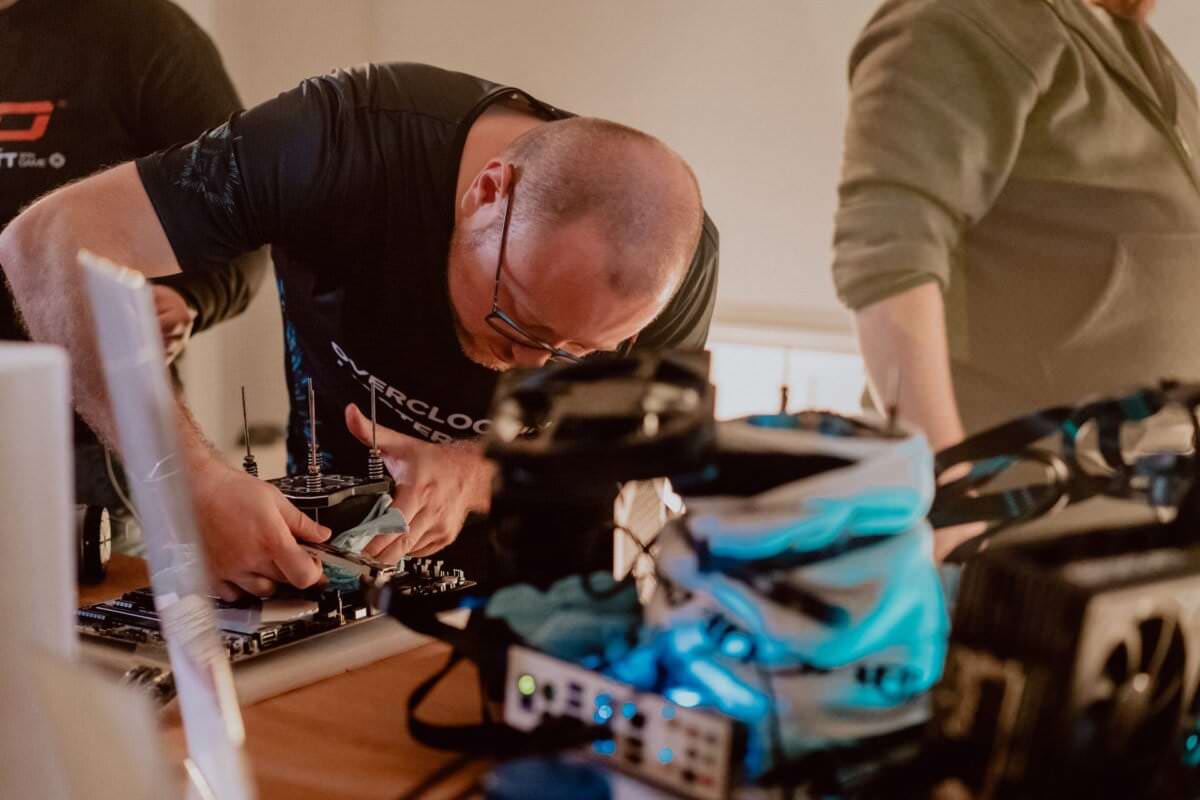 A person with a shaved head and wearing glasses is leaning over a table, carefully working with a tool on a circuit board. The person is wearing a black T-shirt with text on it. Nearby stands another person wearing a green top. Various electronic components are scattered on the table to capture the perfect photo of the event.   
