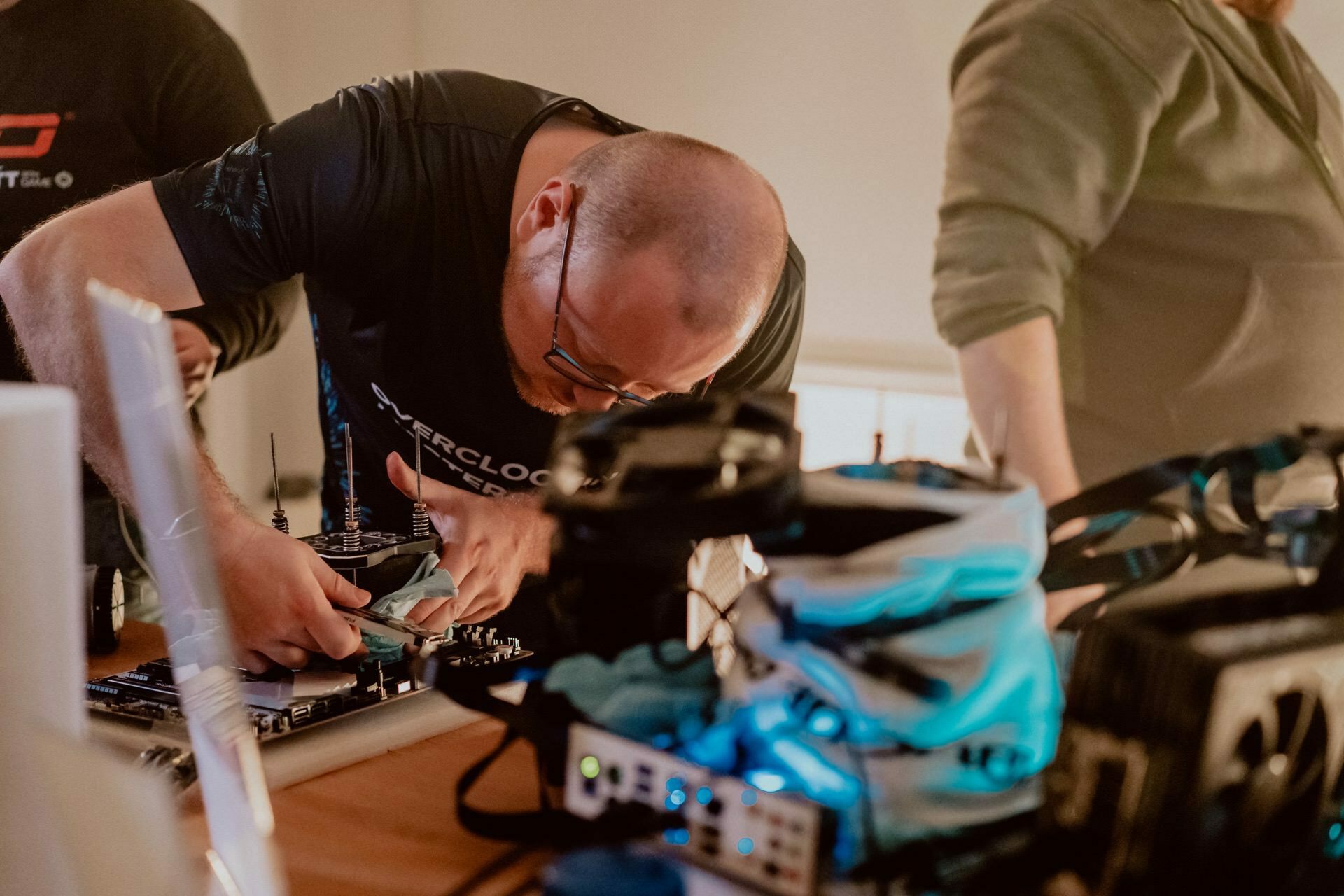 A person with a shaved head and wearing glasses is leaning over a table, carefully working with a tool on a circuit board. The person is wearing a black T-shirt with text on it. Nearby stands another person wearing a green top. Various electronic components are scattered on the table to capture the perfect photo of the event.   