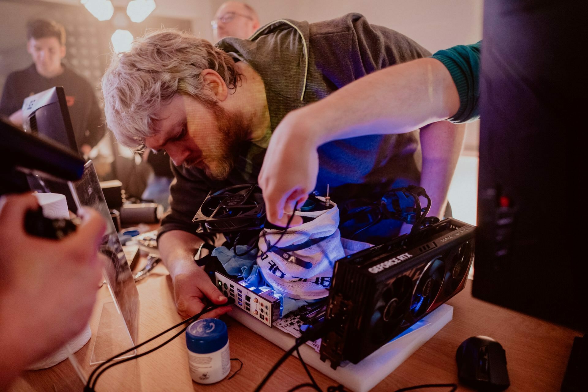 A person with gray hair and beard is busily working on the computer's internal components on a desk, while several people watch in the background. The set includes various tools, cables and a large graphics card. Blue lighting illuminates the scene, capturing a captivating photo essay of the event.  