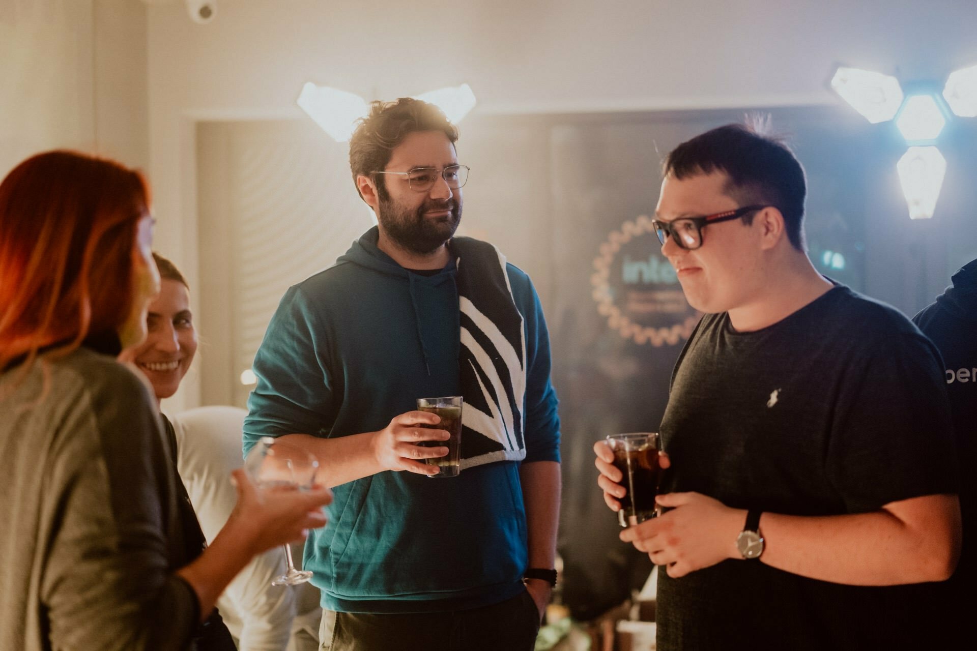 A group of four people are meeting in a house, having drinks. Two people are busy talking and smiling. The background has warm lighting with visible decorative lights that create a cozy atmosphere. The essence of Marcin Krokowski's event photography is reflected in this scene.   