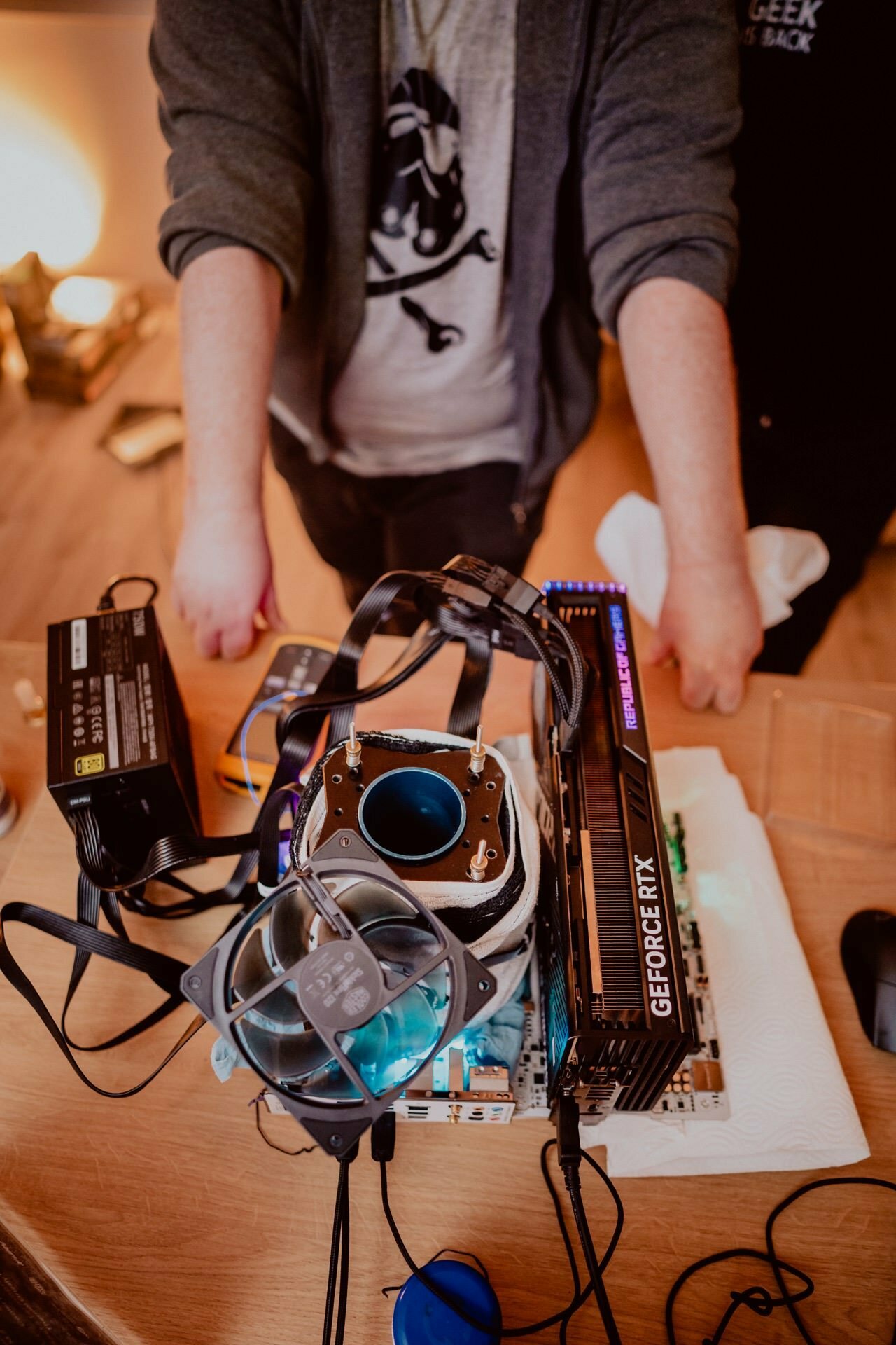 A person is working on building a computer on a wooden table. Components, including a GeForce RTX graphics card, CPU cooler and other parts, are scattered about. A person wearing a jacket with a skeleton graphic has his arms stretched out over the table. Event photographer Marcin Krokowski captured the scene perfectly.   