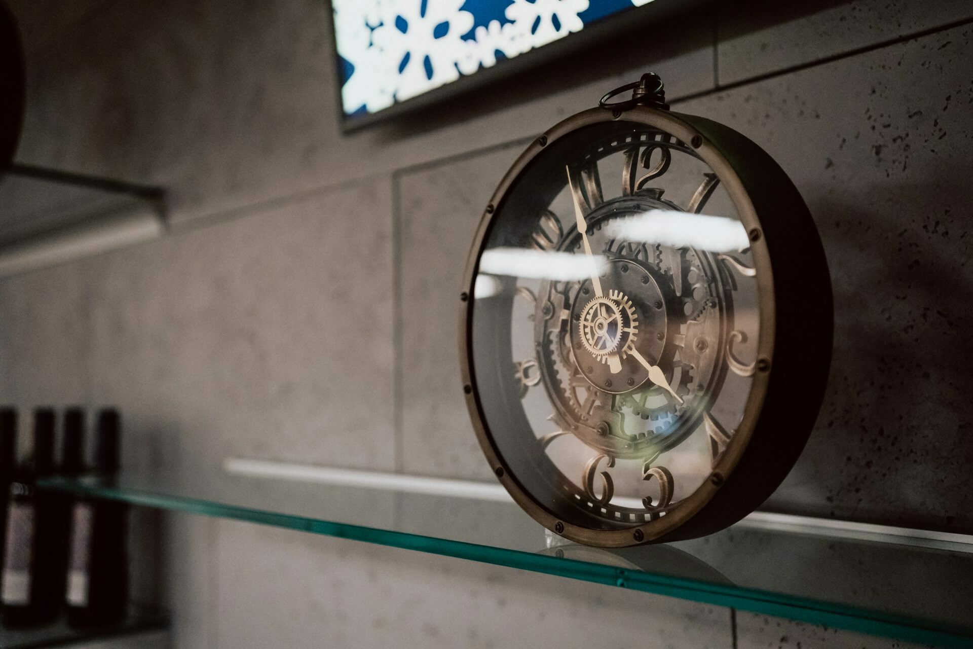 An ornate bronze clock with visible gears stands on a glass shelf. In the background is a tiled wall, and the lower glass shelf shows part of two bottles. The clock is kept in a steampunk style, perfect for a photo essay of events by marcin krokowski.  