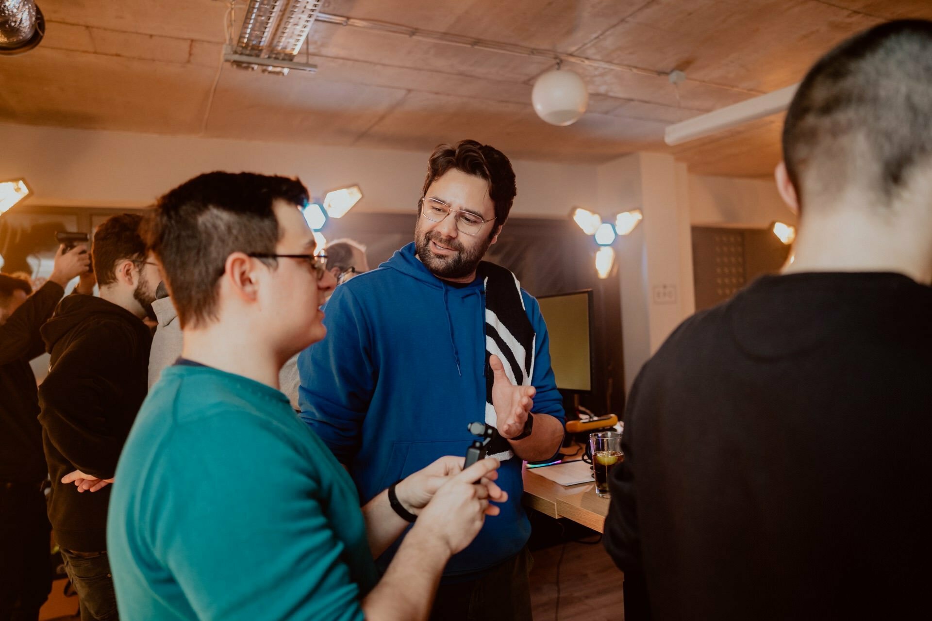 Two people are talking during a social event taking place in a room. One person in a blue sweatshirt gestures while talking to another person in a green shirt holding an object. Several other people mingle in the warmly lit room, as captured perfectly by the event photography.  