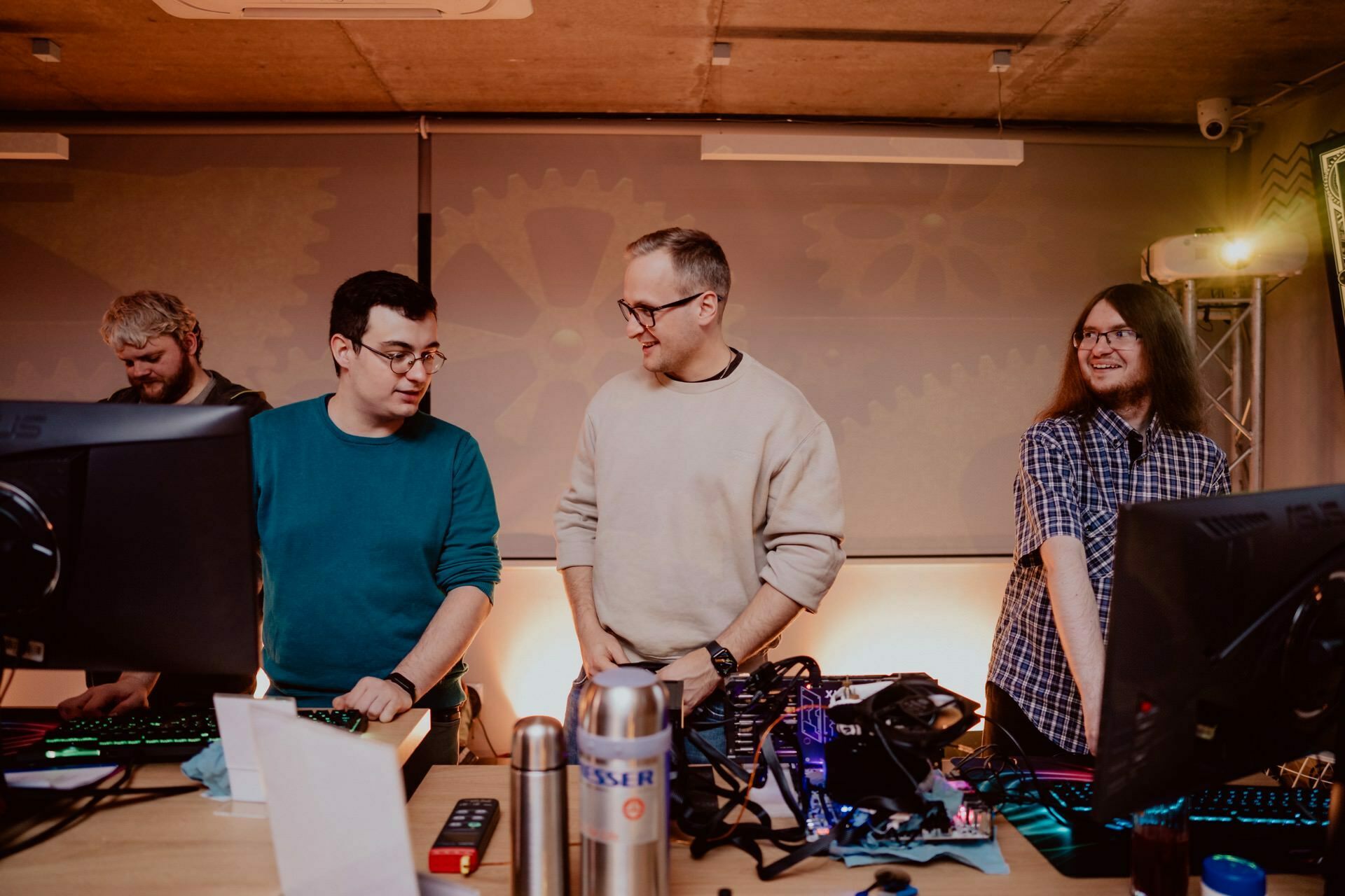 Four people stand at a table with computers and various electronic equipment. Two in the middle are busy talking, while the others on either side are focused on their tasks. In the background is an industrial decor with gear-like motifs, perfectly captured by an event photographer Warsaw for event photo coverage.  