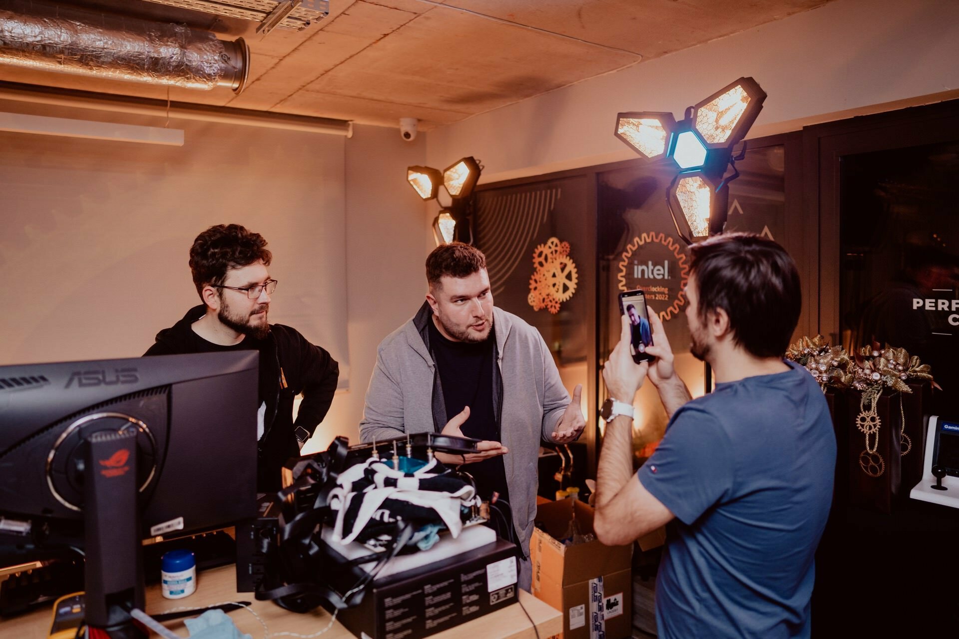 Three people have gathered in a room with technical equipment. Two stand behind a desk with a monitor; one holds an object and gestures. The third person captures the moment with a smartphone, highlighting the dynamics of the event, which is perfect for a photo essay of the event. The room has a warm, industrial atmosphere.   