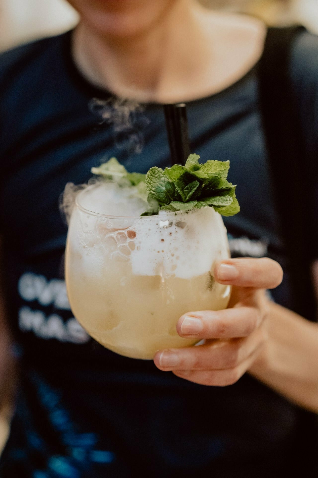 A person holding a round glass filled with a frothy light-colored drink decorated with fresh mint leaves. The drink is sprinkled with a few ice cubes and comes with a black straw. In this photo of the event captured by Marcin Krokowski, the face and upper body of the figure are slightly blurred in the background.  