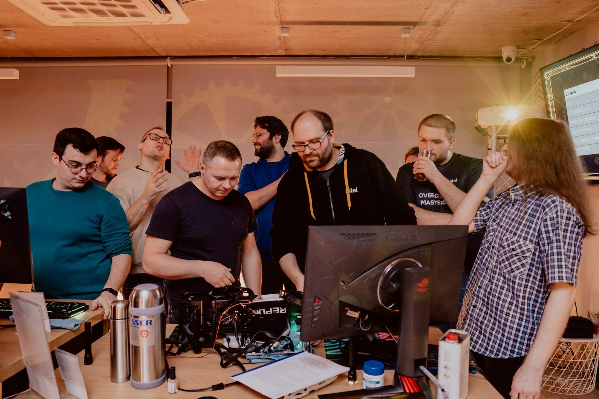 A group of people have gathered around a table with computer equipment and monitors, engaged in technology-related activities. Some focus on the screens, while others interact and discuss. The atmosphere, ideal for event photography, seems collaborative and busy.  