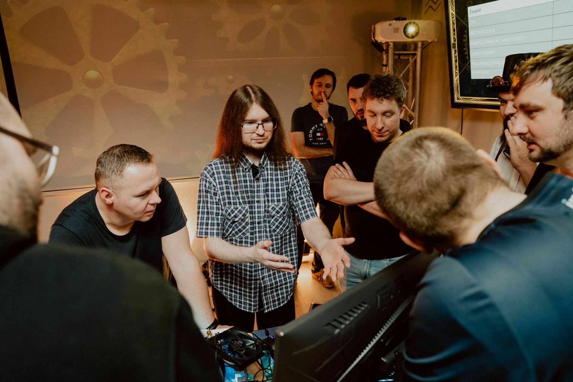 A group of people gather around a table focusing on a project. One person speaks and gestures with their hands, while another listens intently. In the background is a screen displaying data and a wall with gear motifs, capturing the dynamic atmosphere, which is documented by Marcin Krokowski, an acclaimed event photographer Warsaw.  