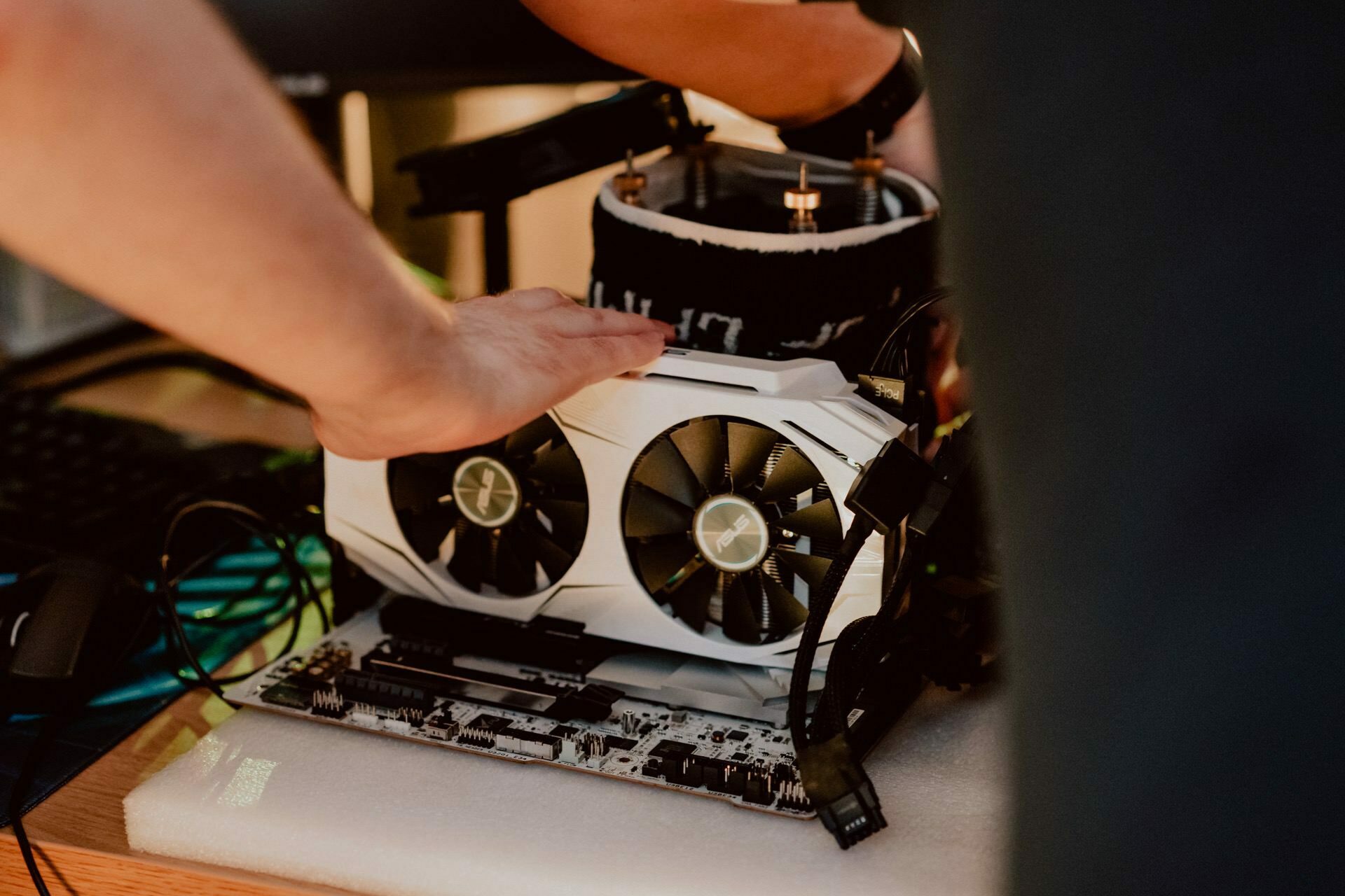 A close-up of the hands of a person working on assembling or modifying a computer. The focus is on the white graphics card with two fans that is mounted on the motherboard. Other computer components and cables are visible in the background, capturing the detailed process often seen in event photos.  