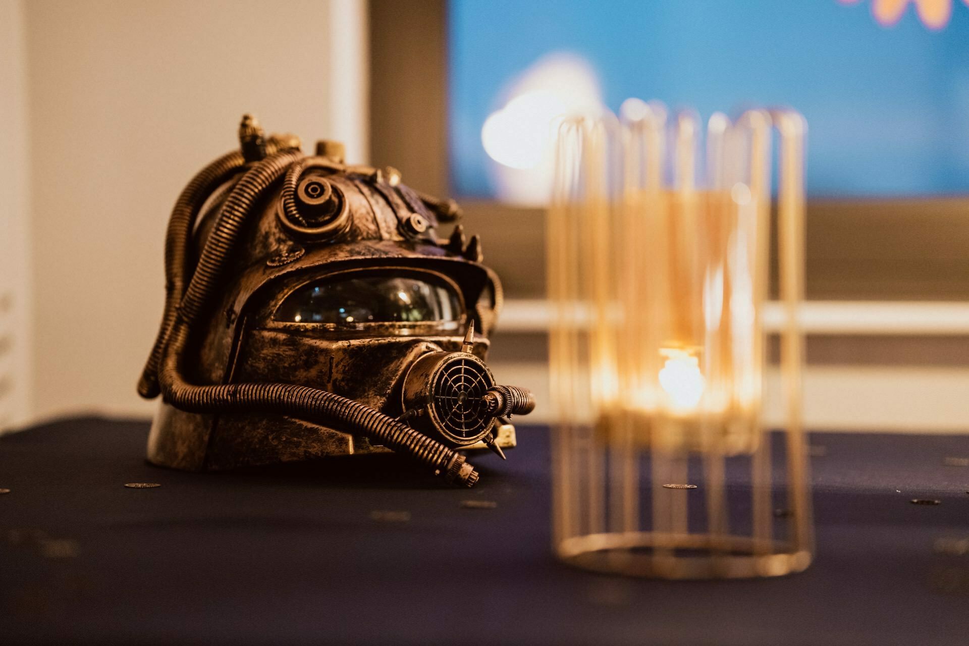 A steampunk-style helmet with intricate metal work, pipes and breathing apparatus lies on a dark blue tablecloth. In the foreground is a cylindrical glass candle holder with soft lighting, creating a warm and moody environment. The photo captures the essence of Marcin Krokowski's unique style in event photography.  