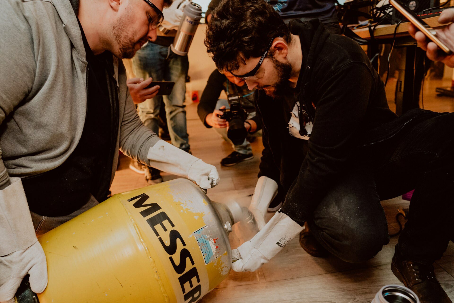 Two men wearing gloves and safety goggles are holding a large yellow container labeled "MESSER," from which gas or steam is probably coming out. Several people in the background are watching and taking pictures, indicating that this may be an organized demonstration or experiment - probably for event photography. 