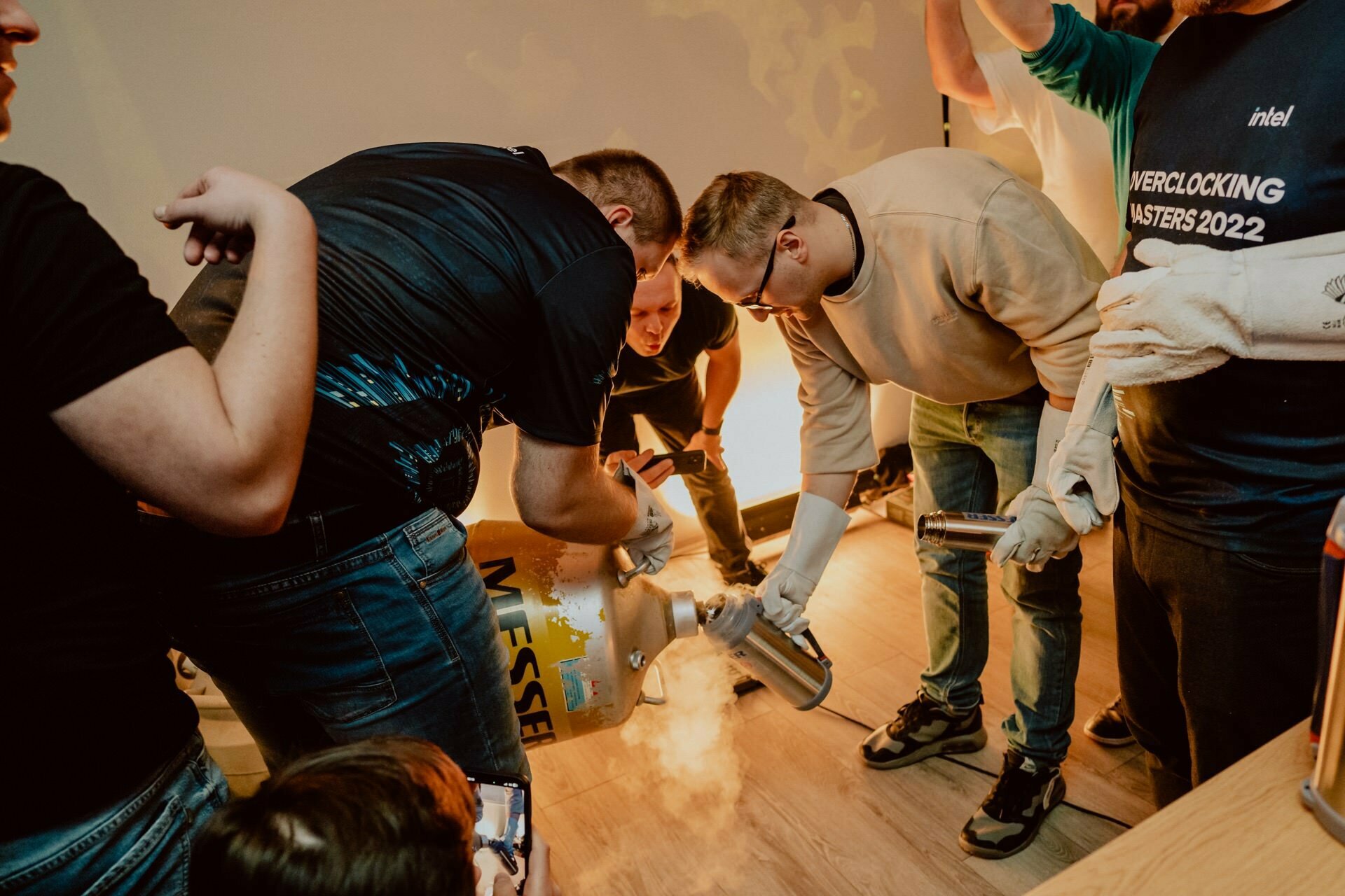 A group of people gather around a large cylindrical container emitting steam. One person pours a liquid from the smaller container, possibly liquid nitrogen, into the larger one. A person in a black shirt holds the cylinder, while someone in a blue shirt looks on. It's intriguing event photography, perfectly capturing the moment.   