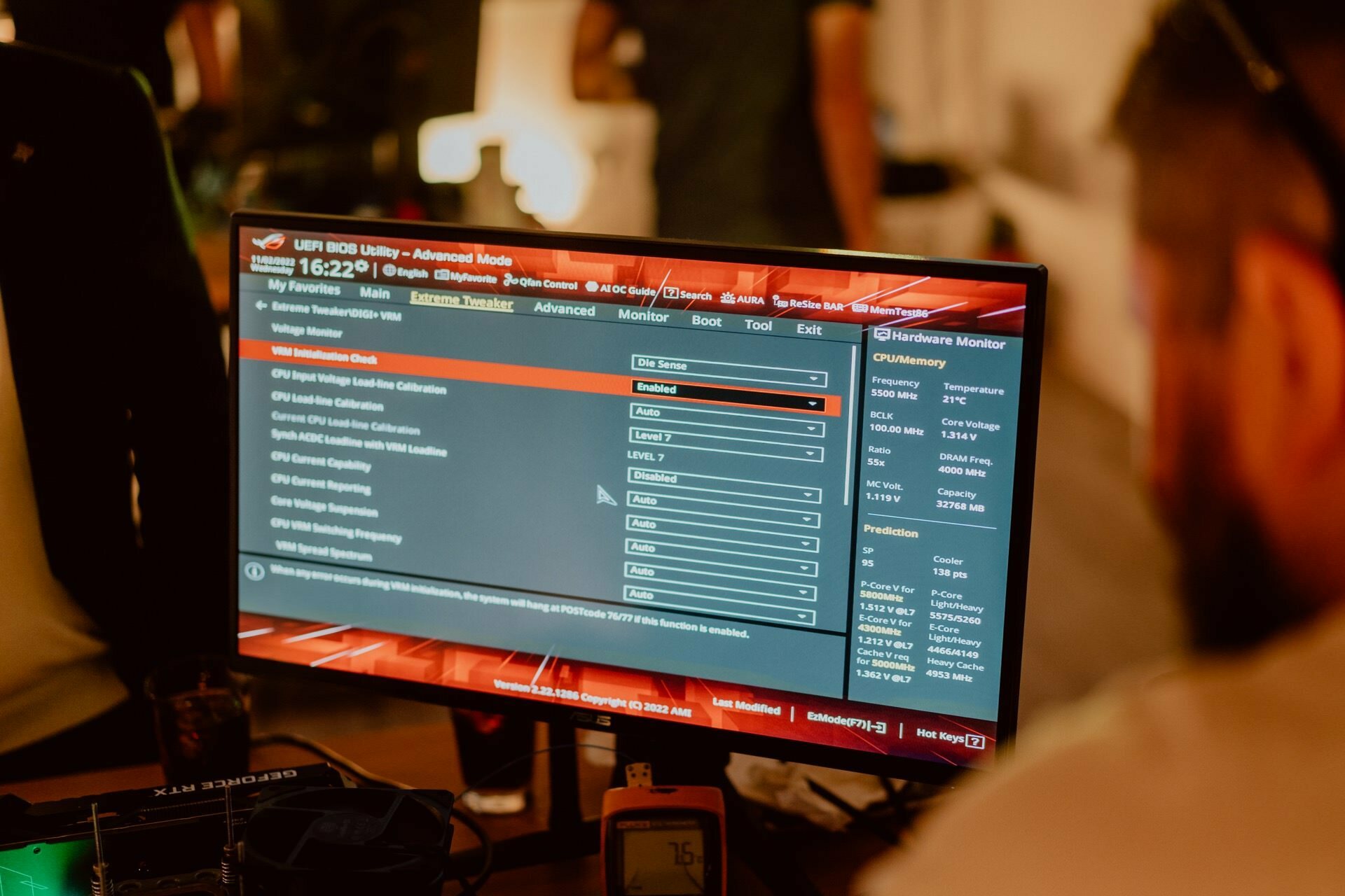 The desktop monitor displays the computer's BIOS interface, showing the processor's configuration settings, including core ratio and other advanced options. In a dimly lit room, the person is partially visible, adjusting the settings. This could be the perfect shot for Marcin Krokowski or any event photographer capturing technology moments.  