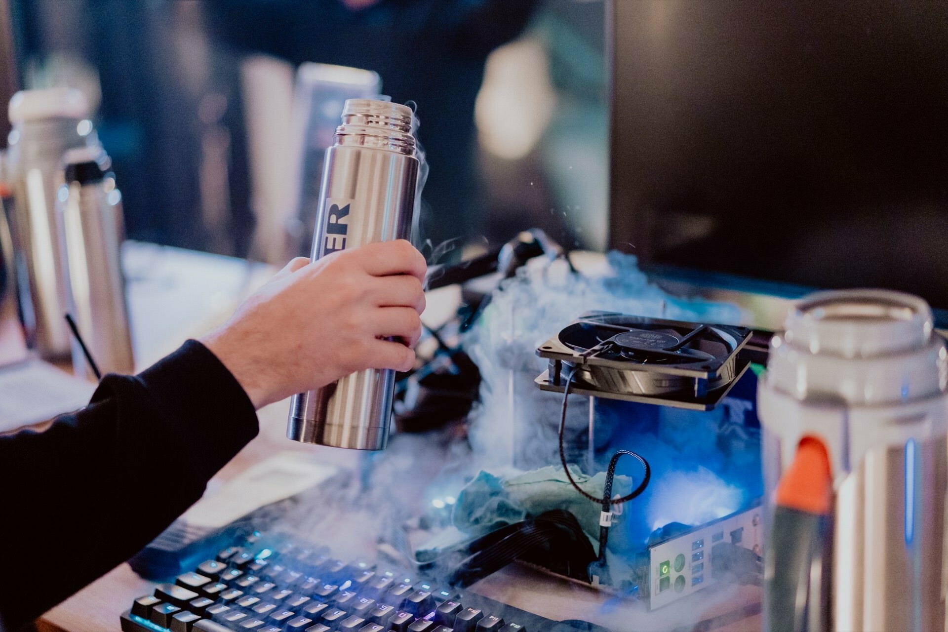 A person holds a metal thermos over a computer showing smoke and ice, suggesting liquid nitrogen cooling while overclocking. The scene captured by Marcin Krokowski consists of a keyboard, a large monitor and multiple containers, indicating a high-tech environment that is likely related to event photography. 