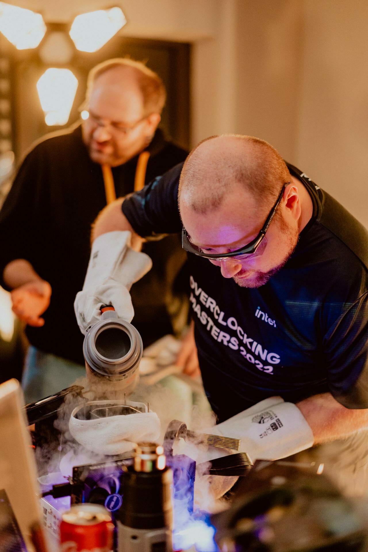 Two people are taking part in an overclocking competition. The person in the foreground, wearing safety goggles and gloves, carefully pours liquid nitrogen into a container. The other person, slightly blurred in the background, watches the process. This scene captures an extraordinary moment for any event photographer Warsaw.   