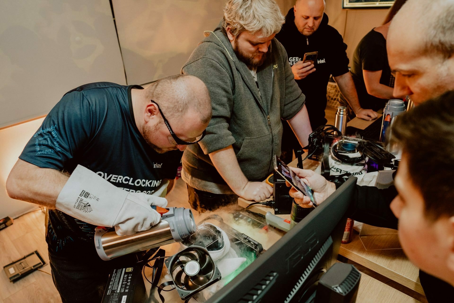 A group of men gather around a table with computer equipment. One of the men, presumably Marcin Krokowski, pours liquid nitrogen into a container while wearing protective gloves, indicating that he is probably engaged in overclocking. In a photo report of the events, various tools and monitors are visible on the table.  