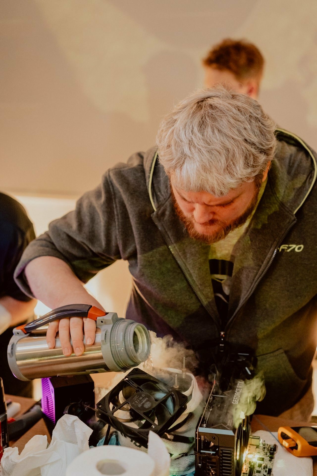 A man with gray hair and beard, wearing a gray jacket, pours liquid from a thermos onto a computer case component. The table is swamped with computer parts and hardware. Marcin Krokowski captures the moment, showing his attention to detail. Another person is blurred in the background.   