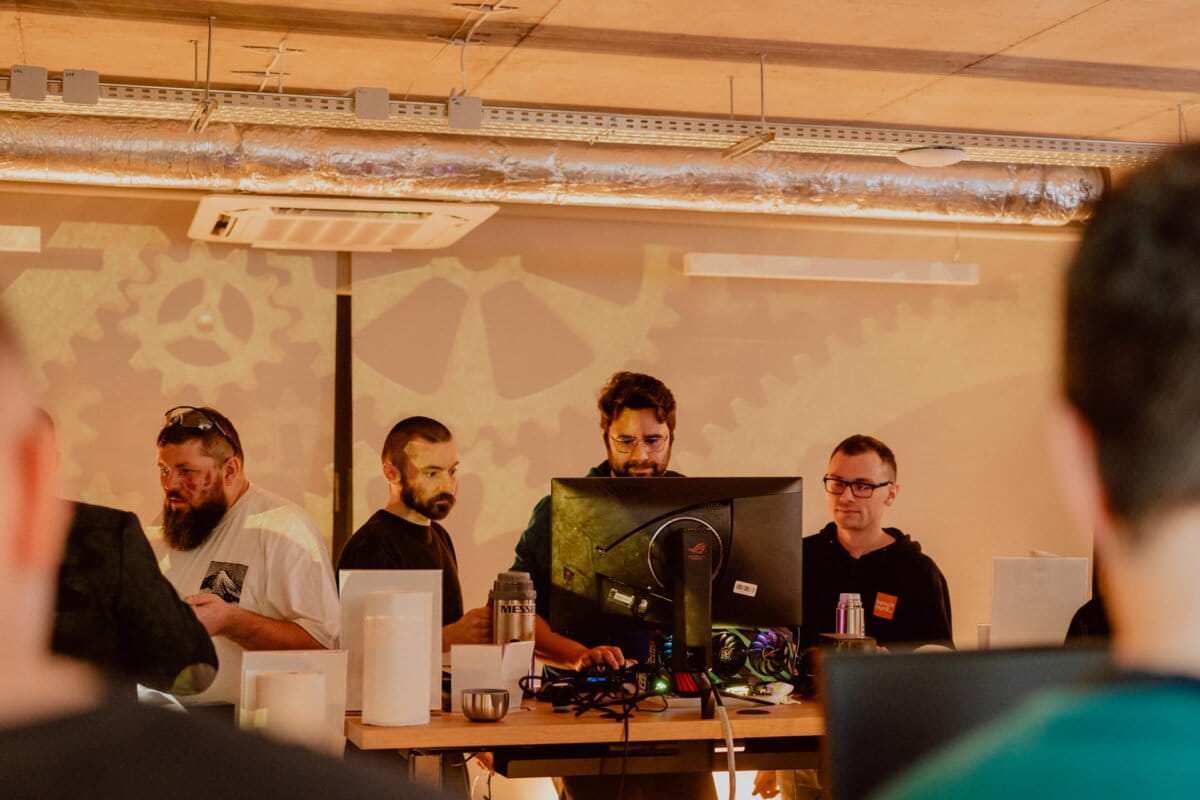 A group of five people are standing around a large computer desk in an office environment. They appear to be collaborating or observing something on the computer screen. The backdrop is industrial, with exposed ducts and a projector displaying cogs on the wall, capturing the atmosphere of a photo-reportage of events.  