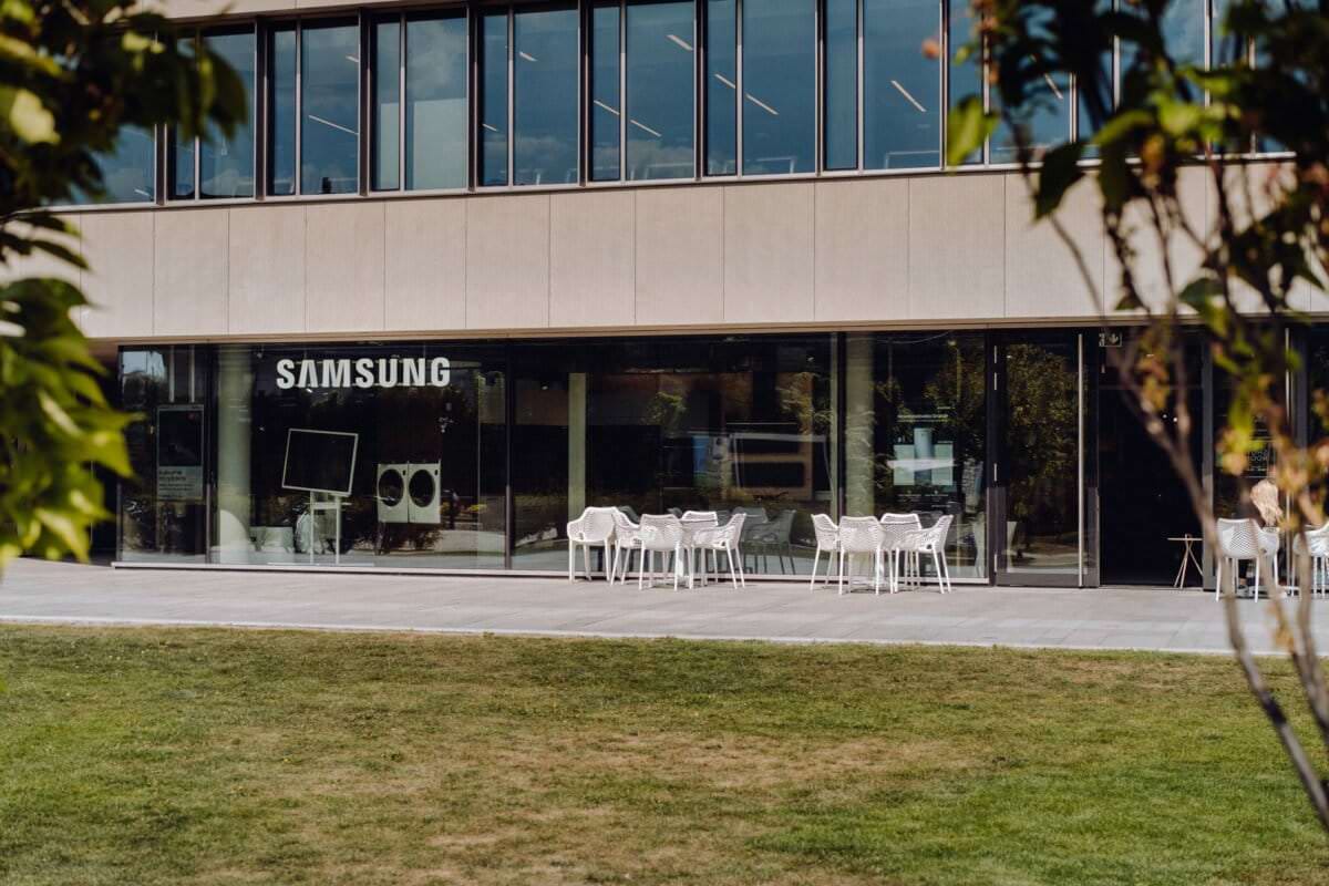 Sklep Samsung ze szklaną fasadą, prezentujący w środku elektronikę. Na zewnątrz na patio znajdują się białe stoły i krzesła, otoczone zielenią i drzewami na pierwszym planie. Budynek posiada duże okna na górnym poziomie – idealne dla fotografa eventowego w Warszawie, który uchwyci tętniącą życiem atmosferę.