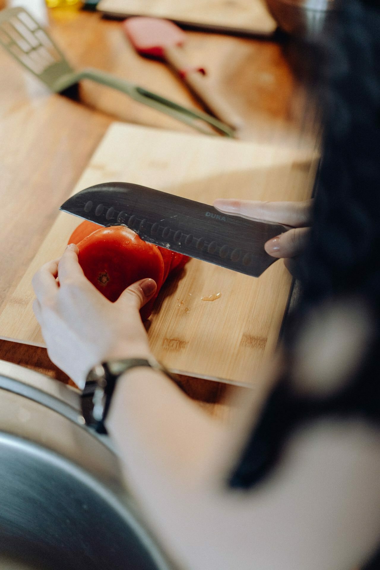 A person with a watch on his wrist is slicing red peppers on a bamboo cutting board using a large kitchen knife. The cutting board stands on a wooden countertop, and kitchen utensils can be seen in the background, creating the perfect setting for event photography. 