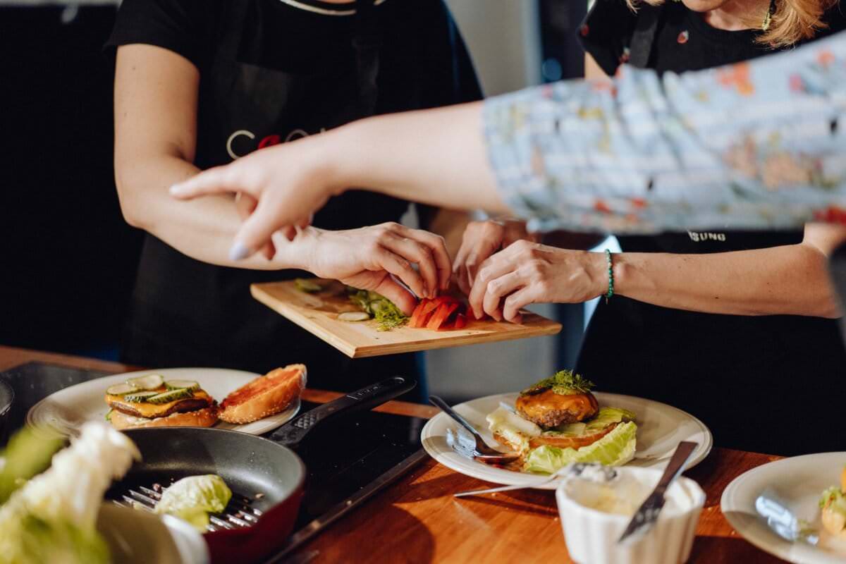 Ludzie gromadzą się wokół kuchennego blatu, składając burgery w ożywionej atmosferze. Jedna osoba trzyma deskę do krojenia z pokrojoną czerwoną papryką, podczas gdy inna układa plasterki na burgerze. Składniki, w tym sałata, są rozrzucone dookoła. Patelnia i talerz stoją na blacie, uchwytując ten idealny moment dla fotografia eventowa.