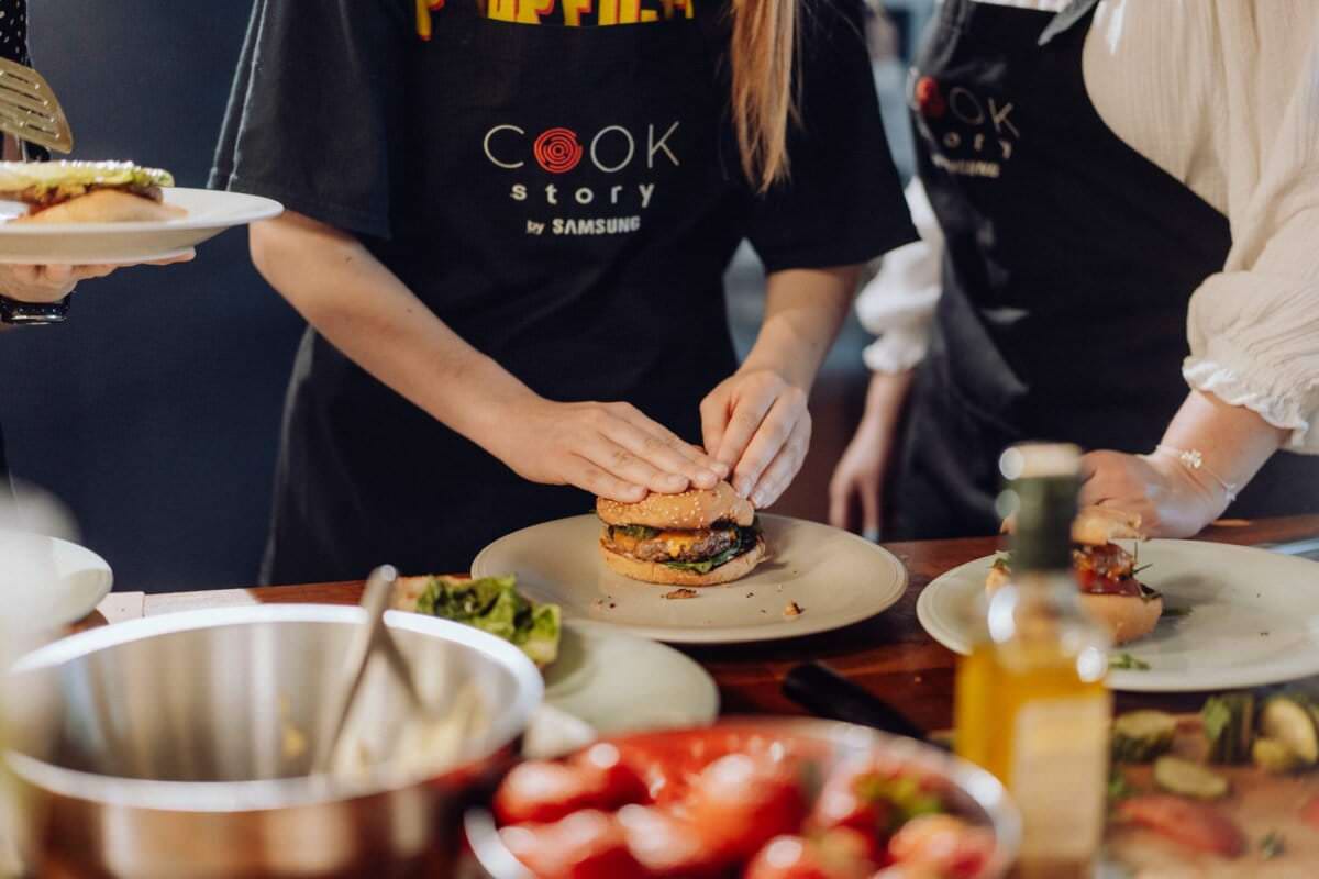 Osoba w czarnym fartuchu z nadrukiem „Cook Story by Samsung” składa burgera na talerzu. Wokół nich na stole rozłożone są różne składniki i dania, w tym miska, sałata, pomidory i butelka oliwy z oliwek. Ten moment oddaje istotę fotografii eventowej w najlepszym wydaniu.