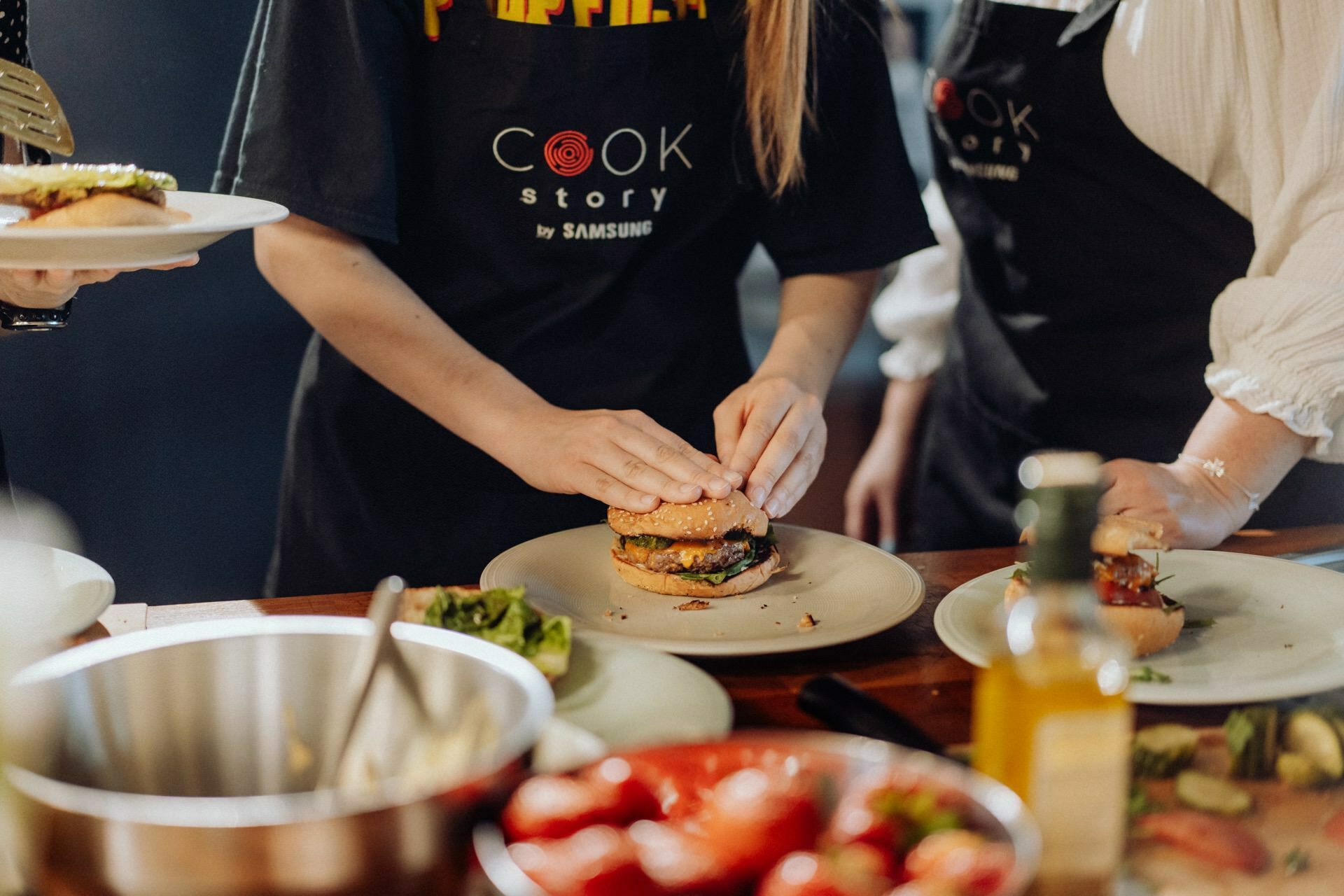 A person wearing a black apron printed with "Cook Story by Samsung" assembles a burger on a plate. Around them, various ingredients and dishes are spread out on a table, including a bowl, lettuce, tomatoes and a bottle of olive oil. This moment captures the essence of event photography at its best.  