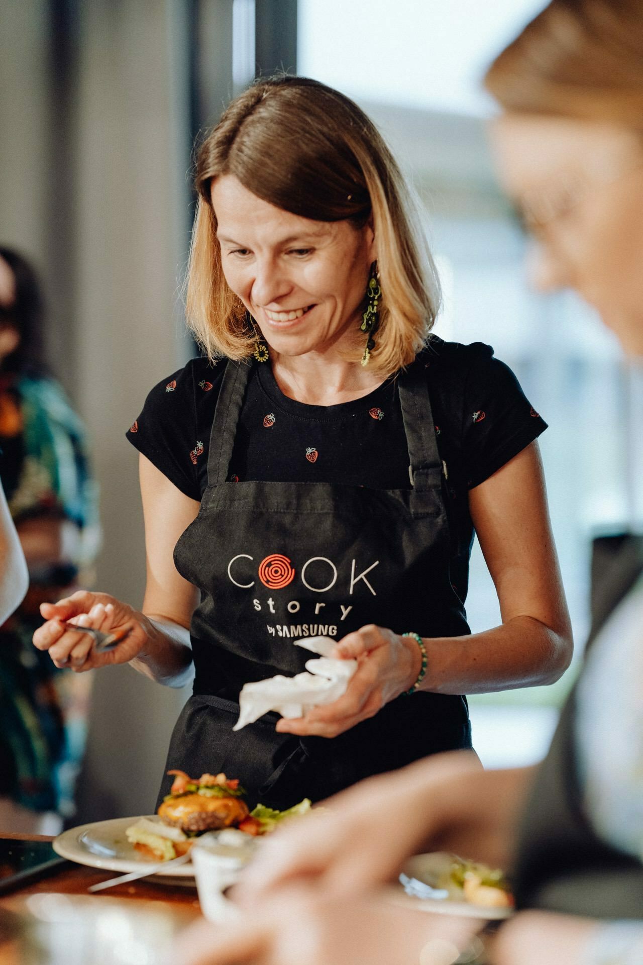 At a cooking event, a woman with shoulder-length brown hair wearing a black apron printed with "Cook Story by Samsung" is preparing food. Smiling and focused on the task, she holds a dish in one hand and a napkin in the other. This photo perfectly captures event photography.  