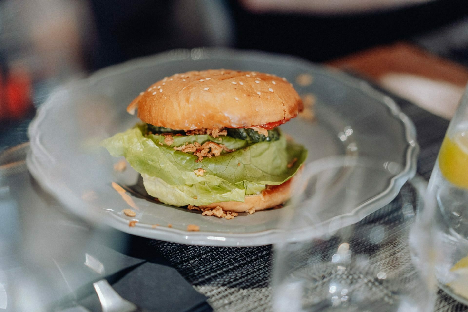 A close-up of a sandwich on a gray plate. The sandwich, perfect for any event photography, consists of shredded lettuce, sliced cucumber and breaded filling, with a sesame seed bun on top. The plate is set on a checkered tablecloth, with a blurry glass in the foreground.  