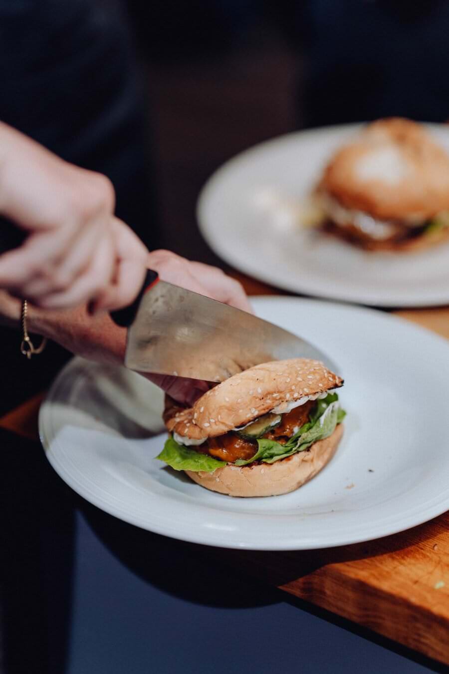 Ręka przecina burgera na pół dużym nożem kuchennym na białym talerzu. Burger ze świeżą sałatą, pomidorem i soczystym kotletem stoi na drewnianej desce. W tle kolejny talerz z podobnym burgerem – idealny do uchwycenia w stylu fotografii eventowej.