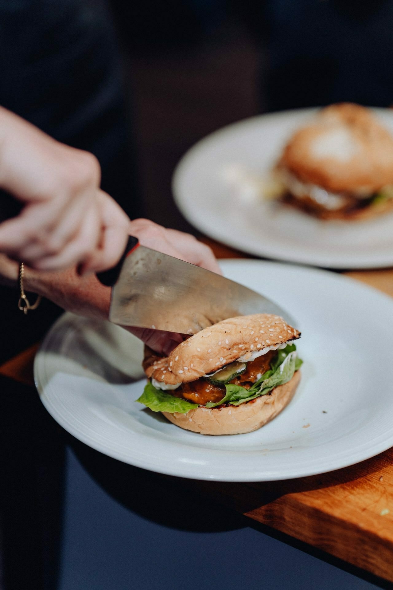 A hand cuts a burger in half with a large kitchen knife on a white plate. The burger with fresh lettuce, tomato and juicy cutlet stands on a wooden board. In the background is another plate with a similar burger - perfect for capturing in event photography style.  