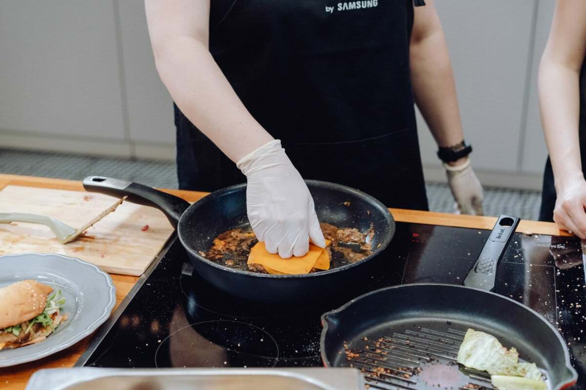 Na tej urzekającej fotografii eventowej osoba ubrana w czarny fartuch i białe rękawiczki trzyma plasterek sera nad skwierczącą patelnią z odrobiną sosu. W pobliżu widać talerz z kanapką i drewnianą deskę do krojenia. Obraz osadzony w nowoczesnej kuchni oddaje istotę kulinarnego rzemiosła.