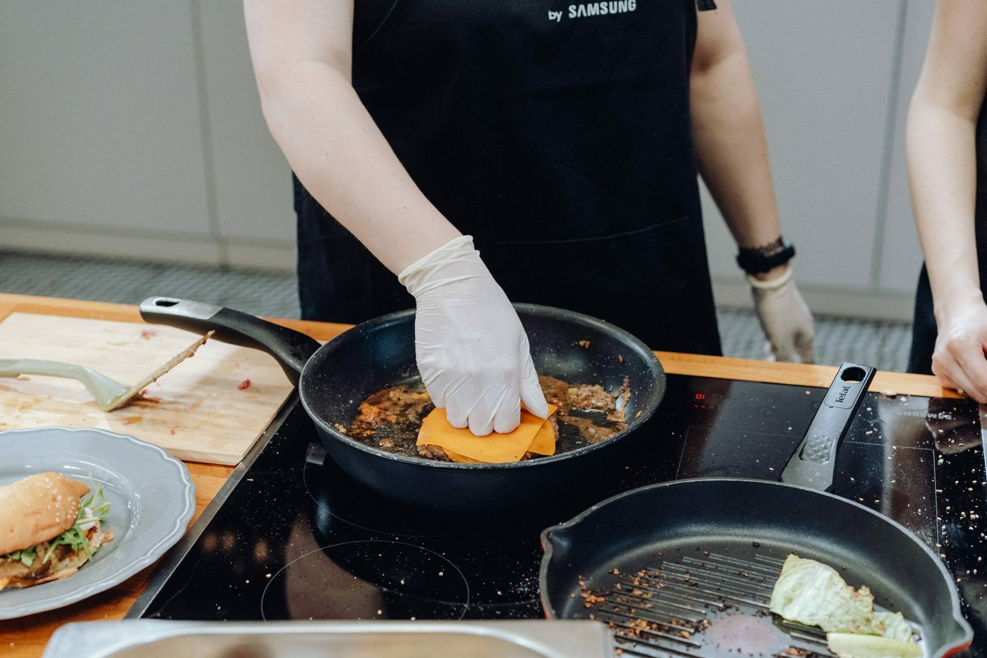 In this captivating event photograph, a person dressed in a black apron and white gloves holds a slice of cheese over a sizzling pan with a bit of sauce. A plate with a sandwich and a wooden cutting board can be seen nearby. Set in a modern kitchen, the image captures the essence of culinary craftsmanship.  