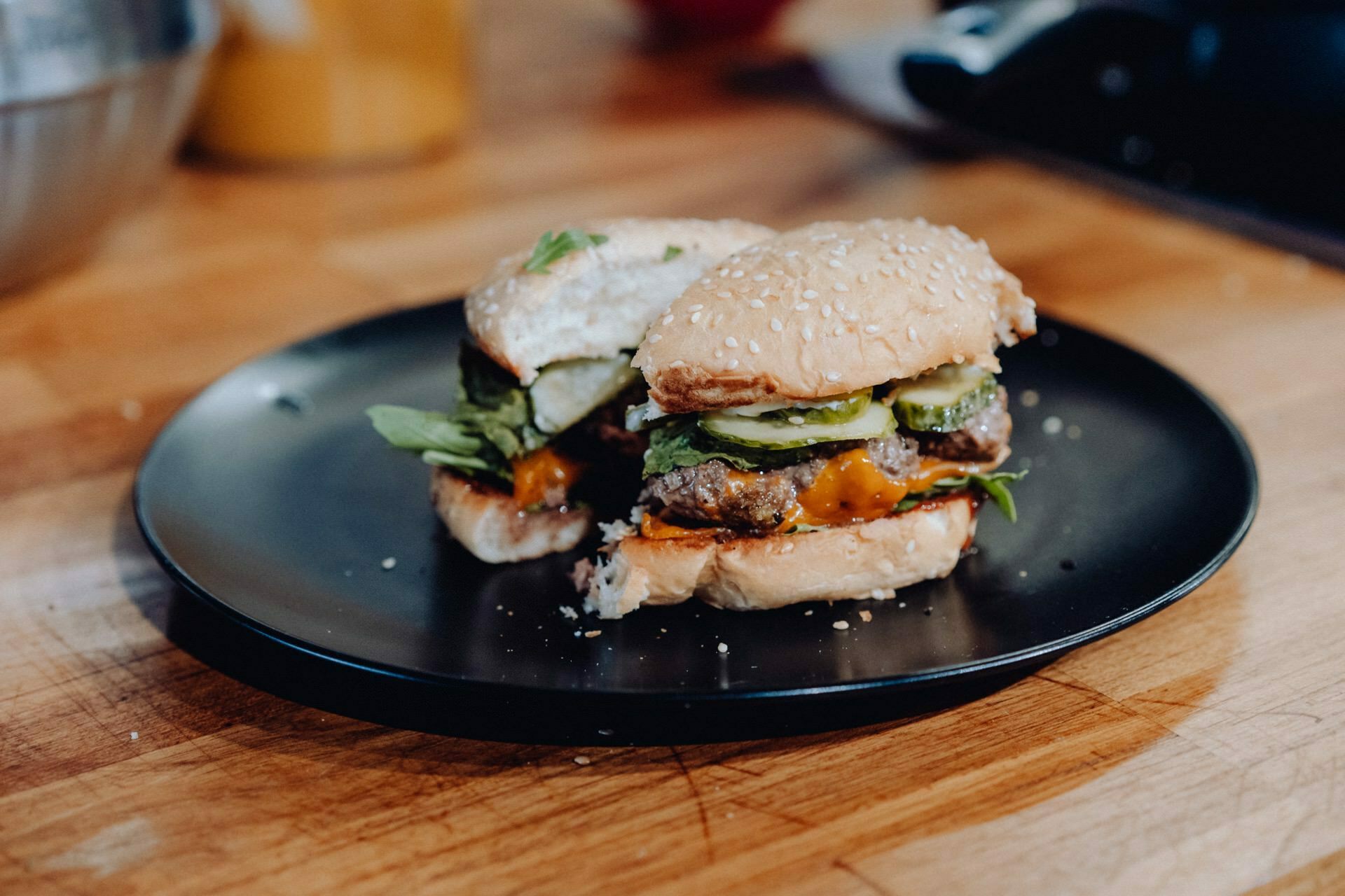 The burger, cut in half, sits on a black plate on a wooden surface. The burger in a sesame seed bun features lettuce, cucumber slices and melted cheese, highlighting its savory ingredients. This photo captures the essence of event photography with a fuzzy kitchen in the background.  