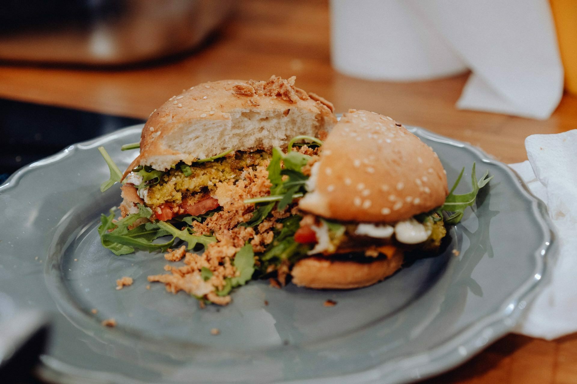 A close-up of a sliced burger on a gray plate. The burger contains lettuce, possibly arugula and other vegetables, and a sesame seed bun. The burger is surrounded by crumbs, and the plate sits on a wooden table with a fuzzy background - perfect for any event photographer of Warsaw to capture the essence of the cuisine.  