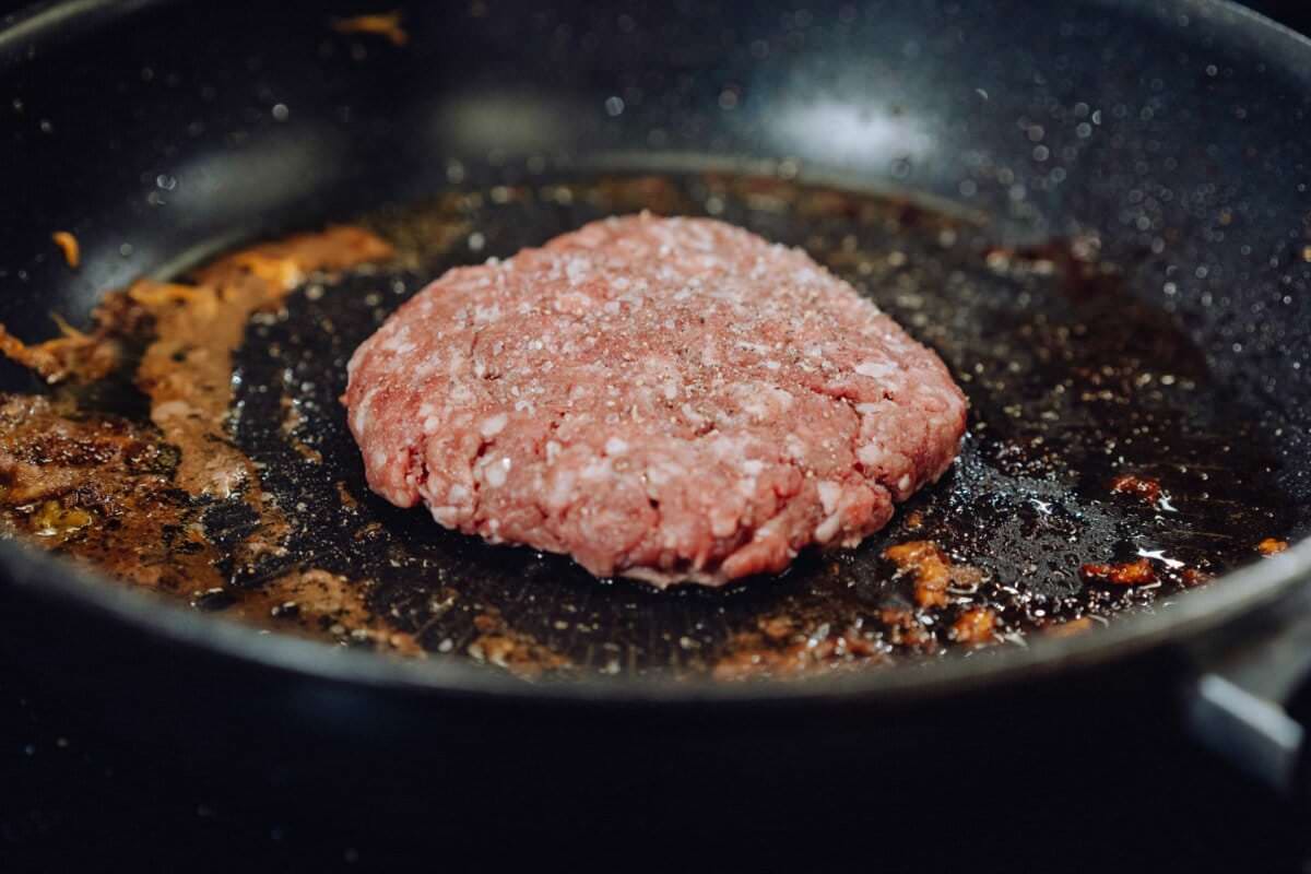 Na czarnej patelni skwierczy surowy kotlet hamburgerowy posypany solą. Patelnia, na której widać resztki oleju i przyrumienione kawałki, oddaje istotę gotowania. Ta scena mogłaby być dla fotografa eventowego Warszawa intrygującym tematem do udokumentowania w fotorelacji z wydarzeń.