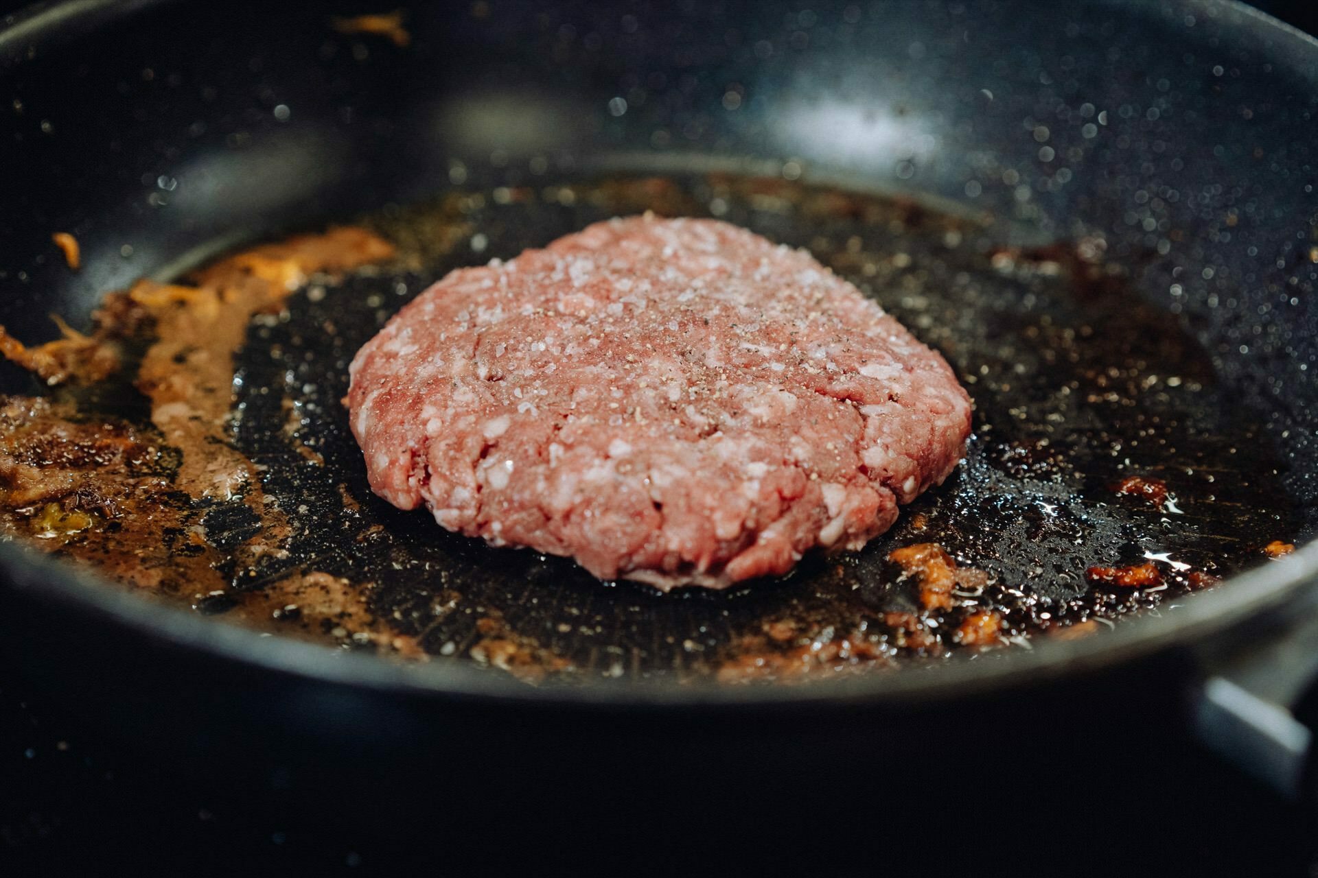 A raw hamburger patty sizzles on a black skillet sprinkled with salt. The skillet, which shows leftover oil and browned bits, captures the essence of cooking. This scene could be an intriguing subject for an event photographer Warsaw to document in an event photo essay.  